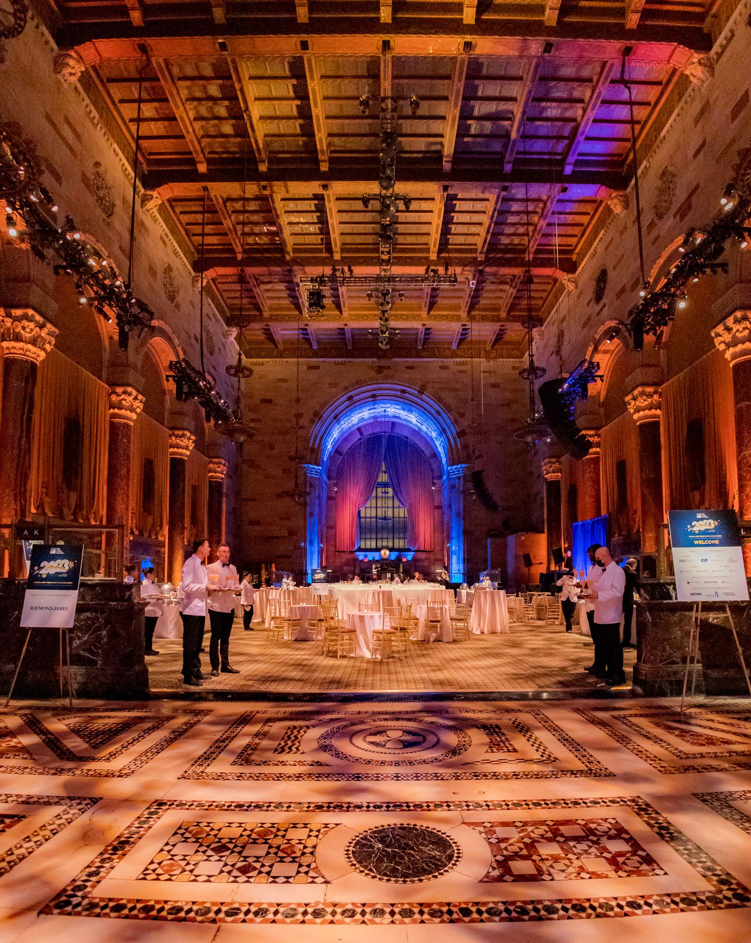Grand ornate event hall with mosaic floor, columns, and blue-lit arched stage.