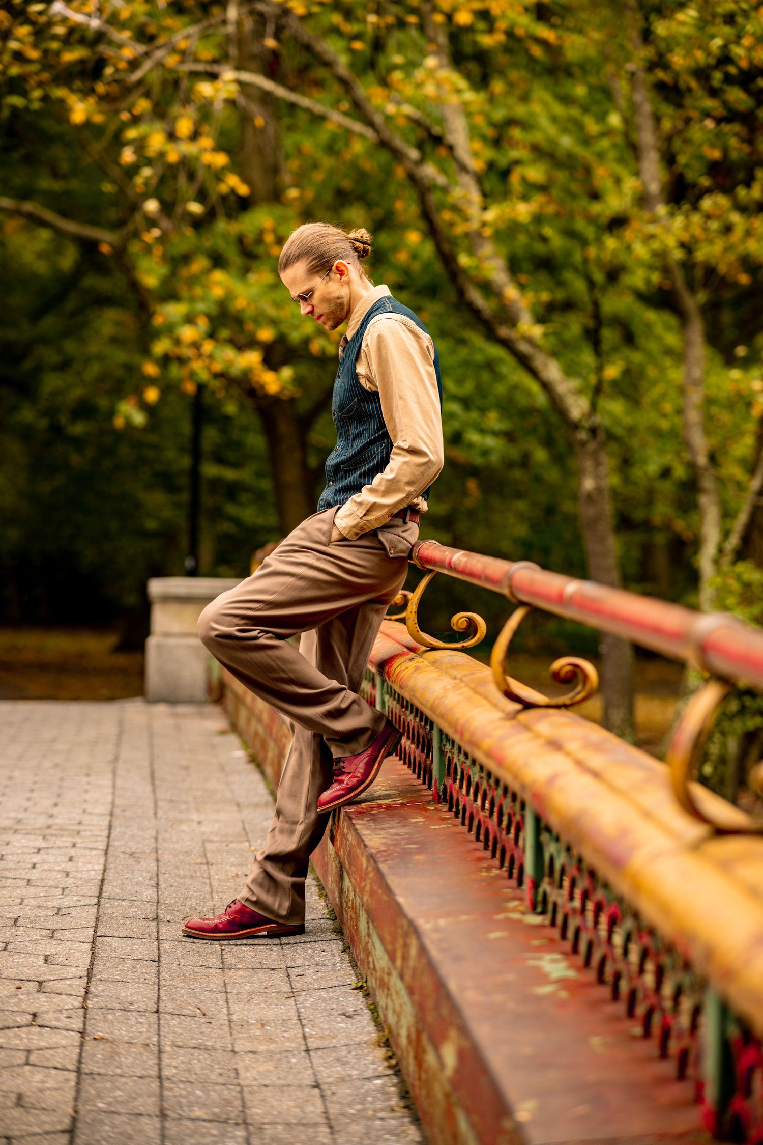 Man in vintage vest and trousers leaning on ornate railing of bridge in autumn park.