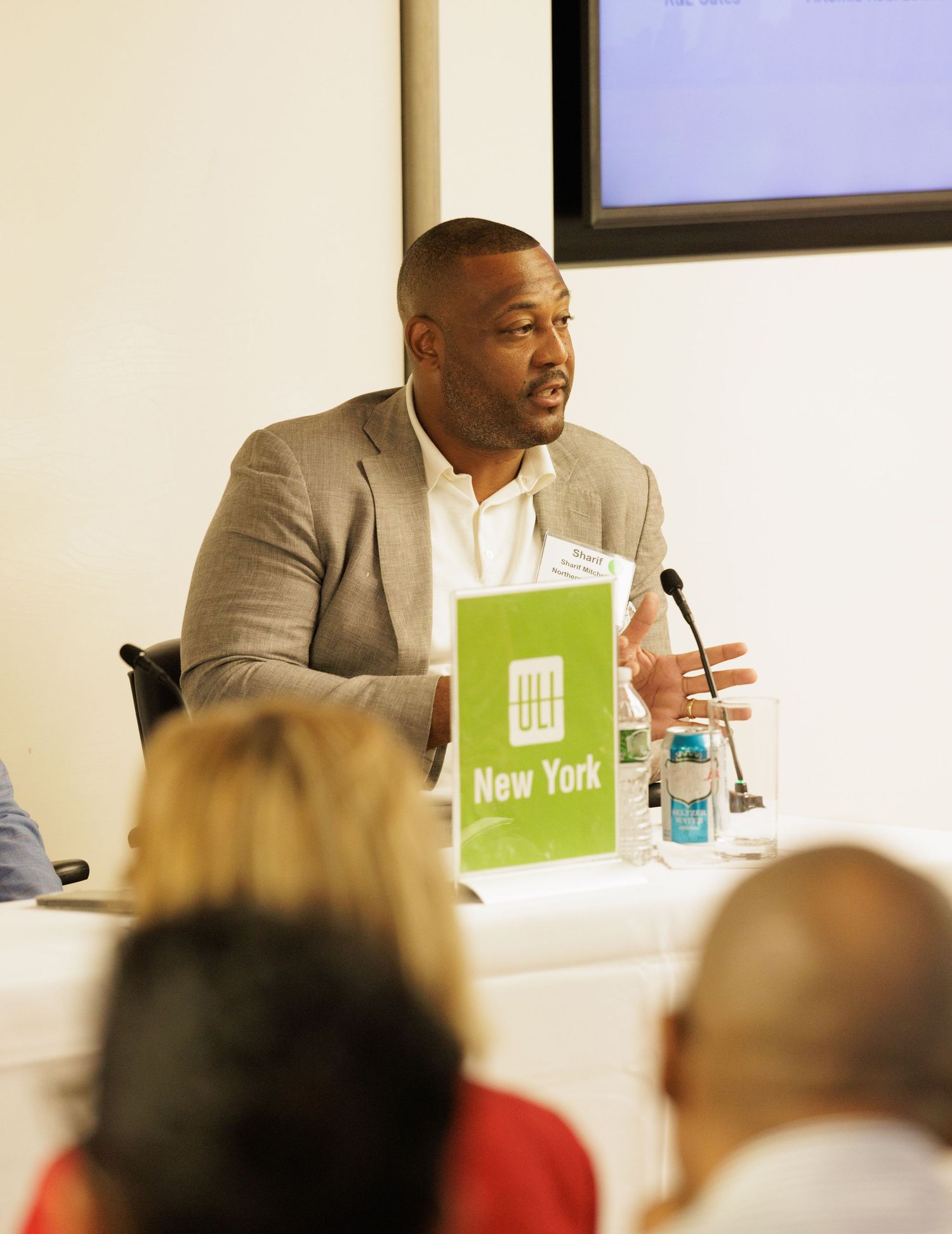 Man in gray blazer speaking at a New York business panel discussion with microphone and green New York sign.