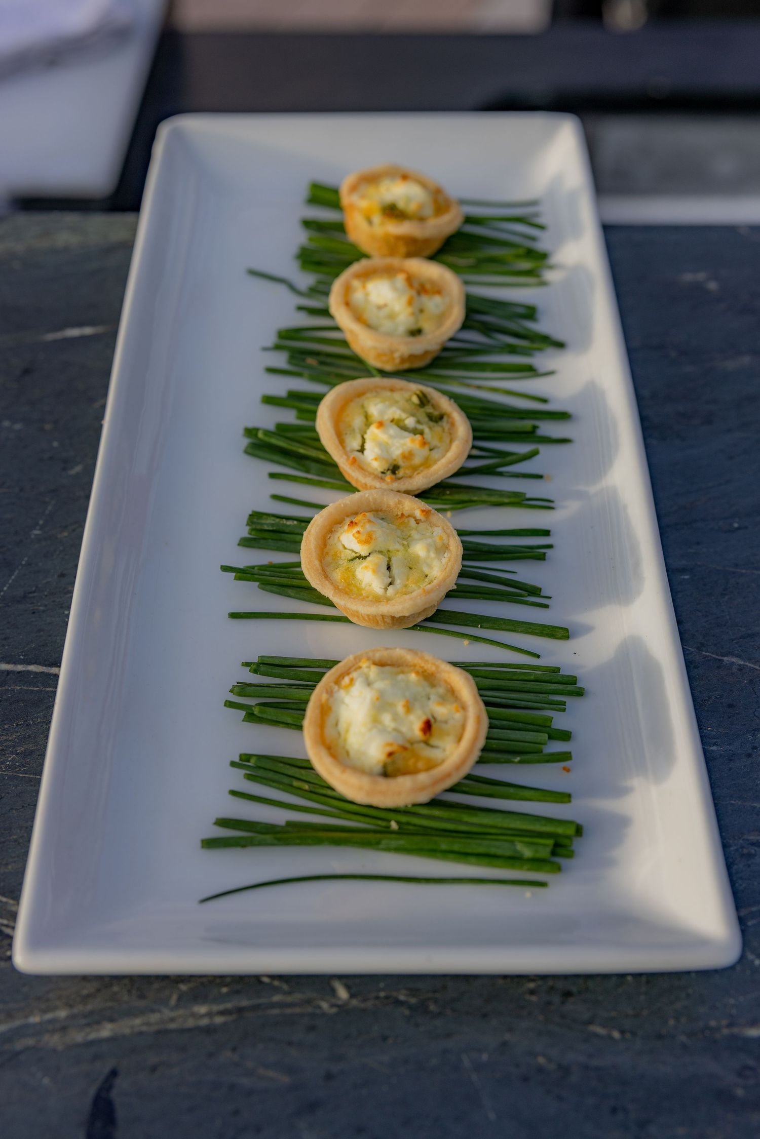 Five mini quiche tartlets arranged on fresh chives on a white rectangular serving plate.