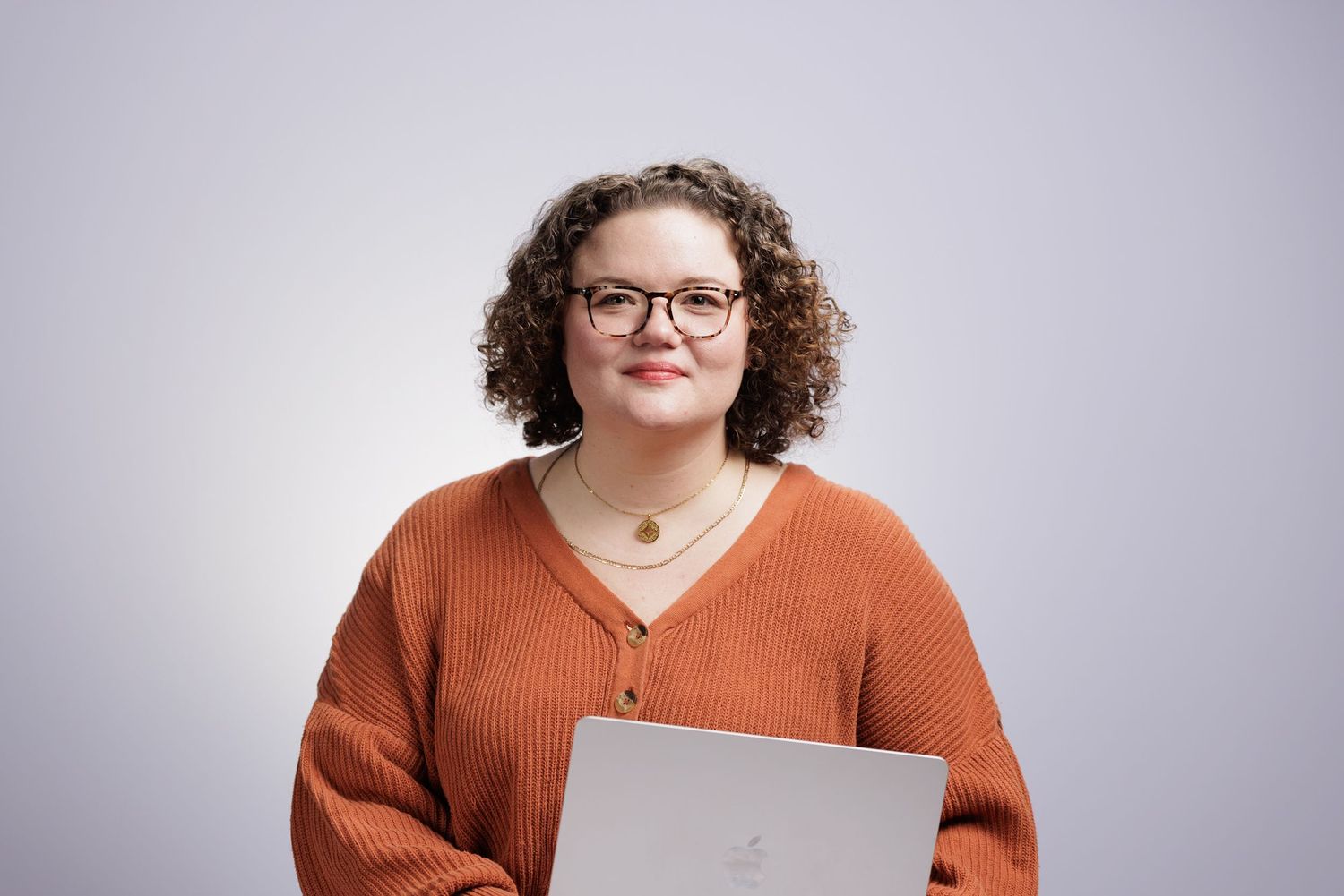 Smiling woman with curly hair and glasses wearing an orange sweater, holding a laptop against a light gray background.