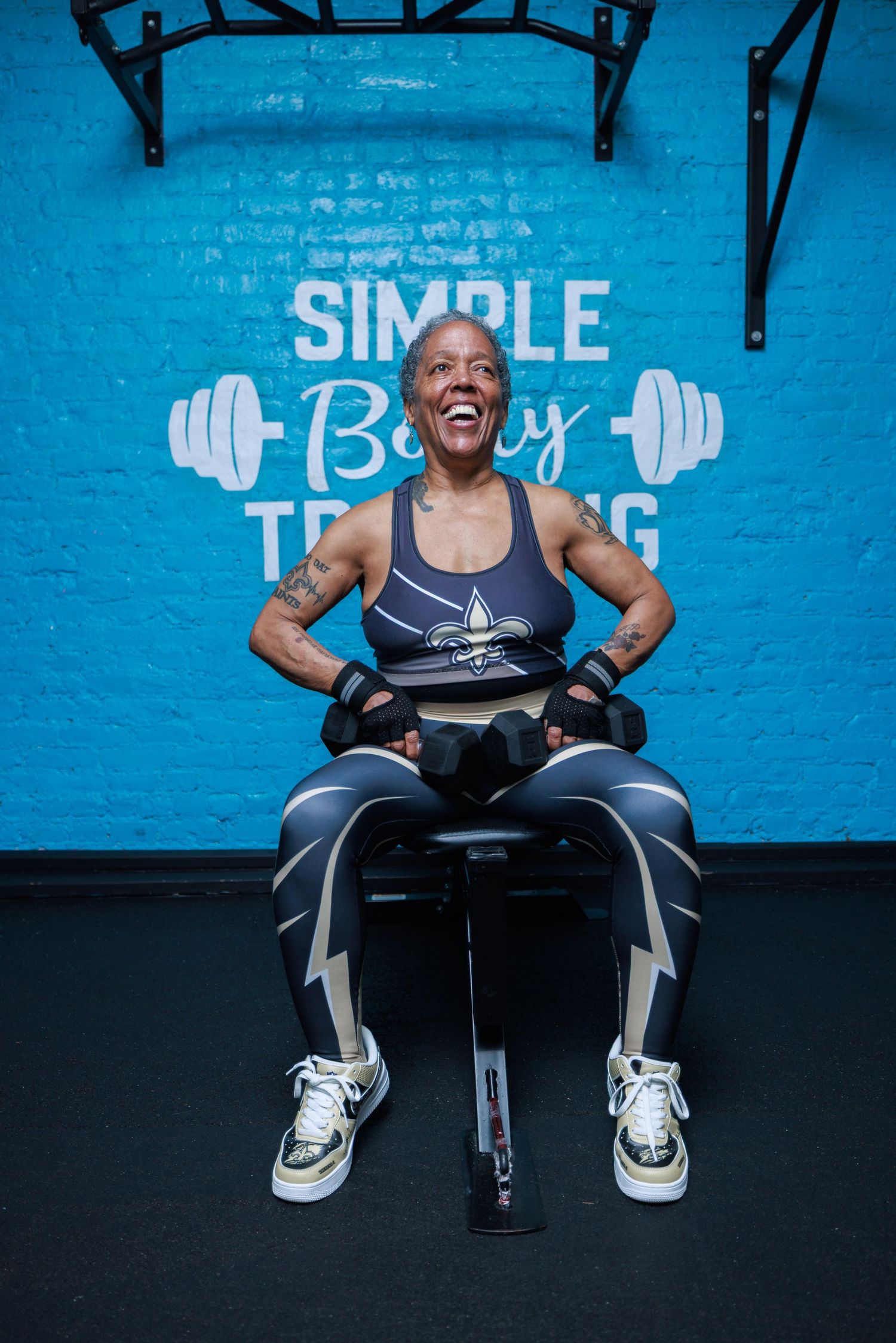 Fit older woman laughing joyfully while seated with dumbbells in a gym with Simple Body Training wall art.