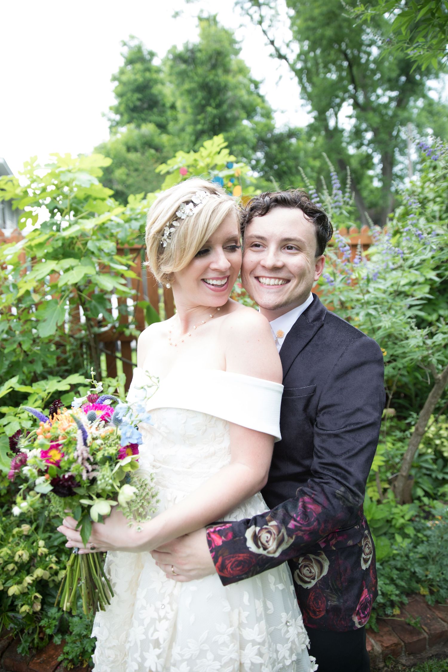 Smiling bride and groom embrace in a lush garden, bride holding a colorful wildflower bouquet in a white lace dress.