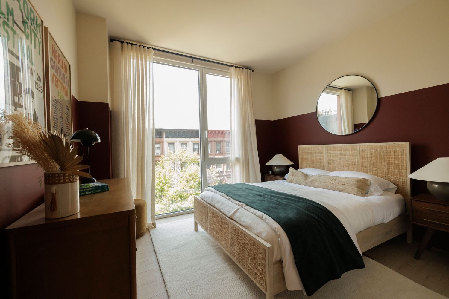 Bright modern bedroom with rattan bed frame, dark accent wall, round mirror, and large balcony window.