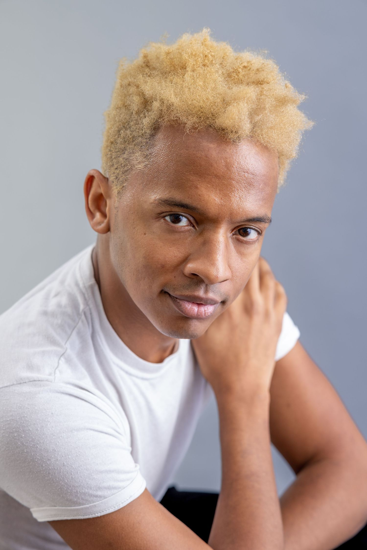 Young Black man with blonde curly hair wearing white t-shirt posing against gray background.