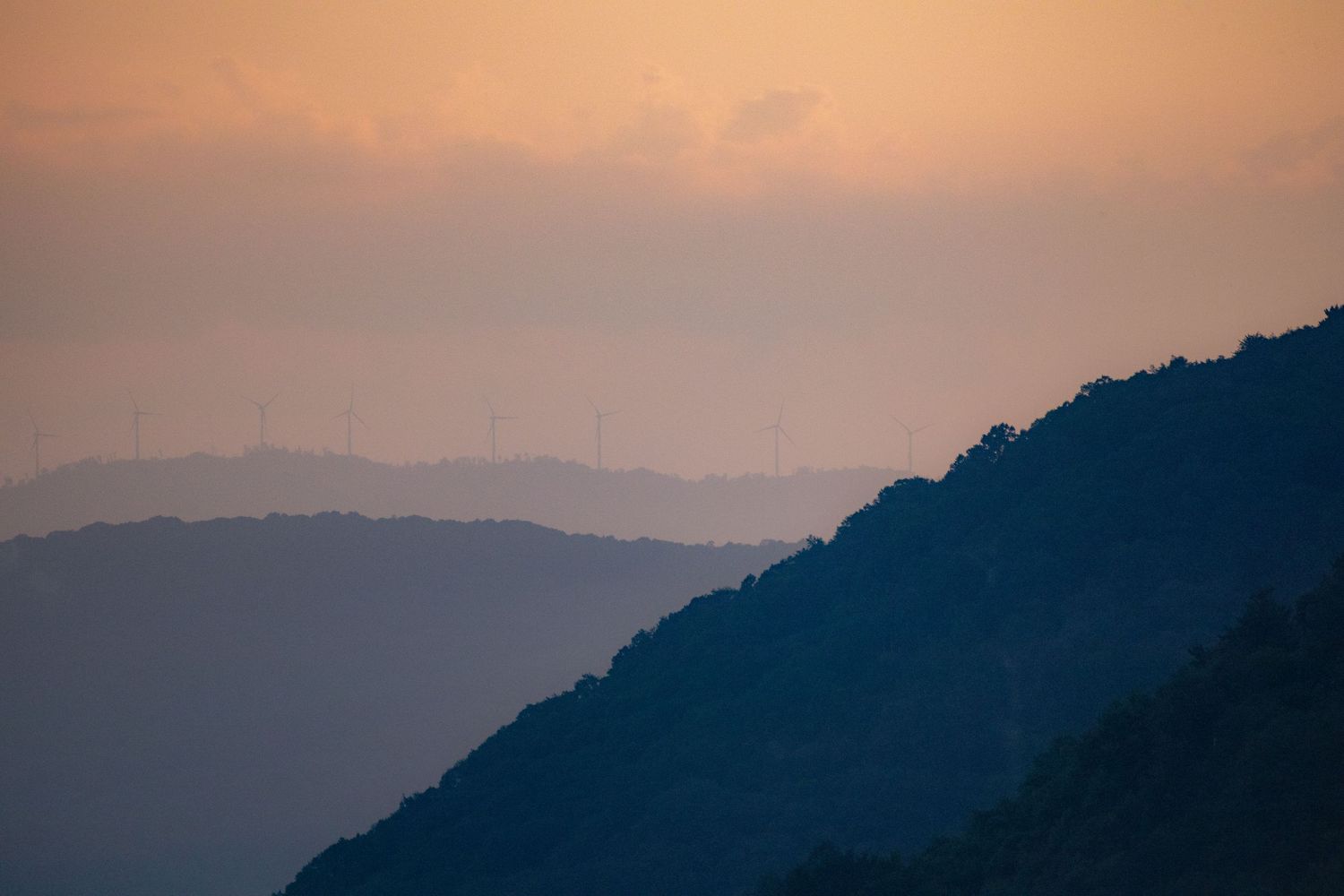 Silhouetted mountain ridges with wind turbines visible through orange hazy sunset sky.