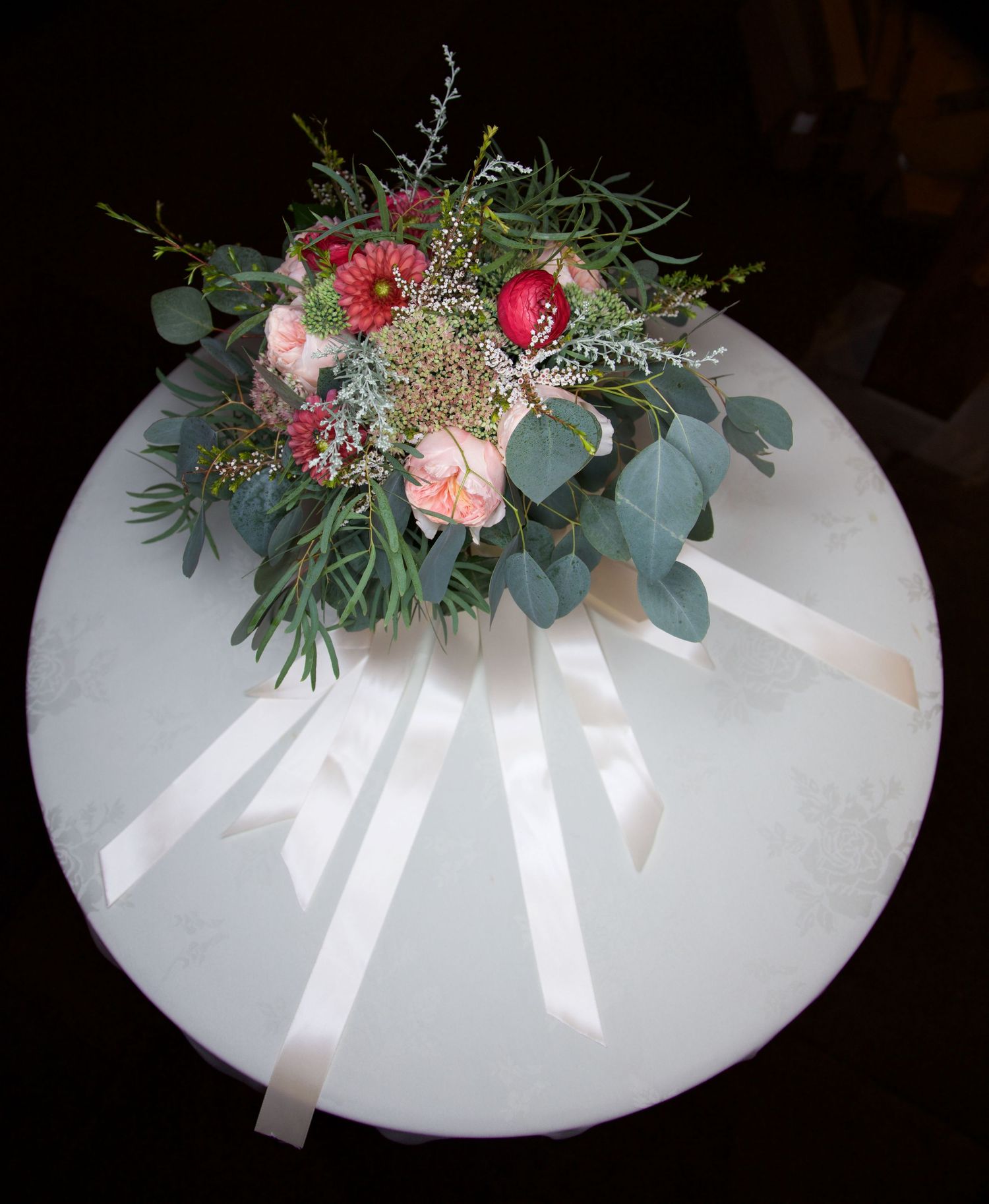 Bridal bouquet with red roses, pink flowers, eucalyptus greenery, and white ribbon streamers on white table.