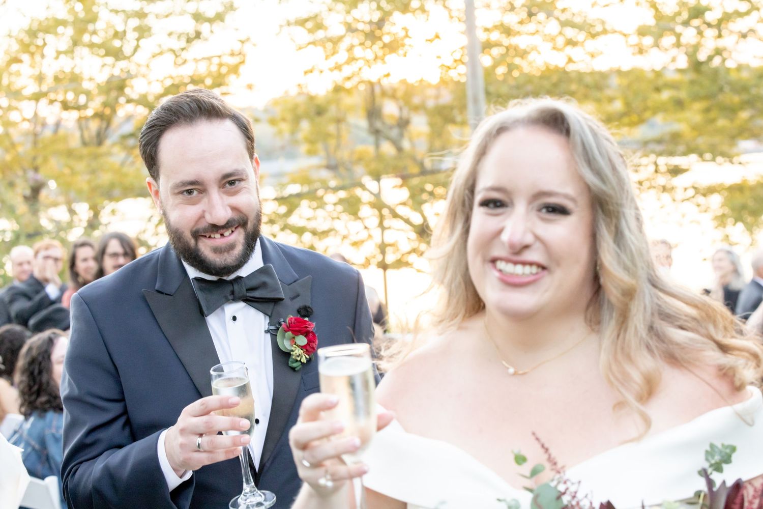 Happy newlyweds toast with champagne glasses at their outdoor wedding celebration with guests behind them.