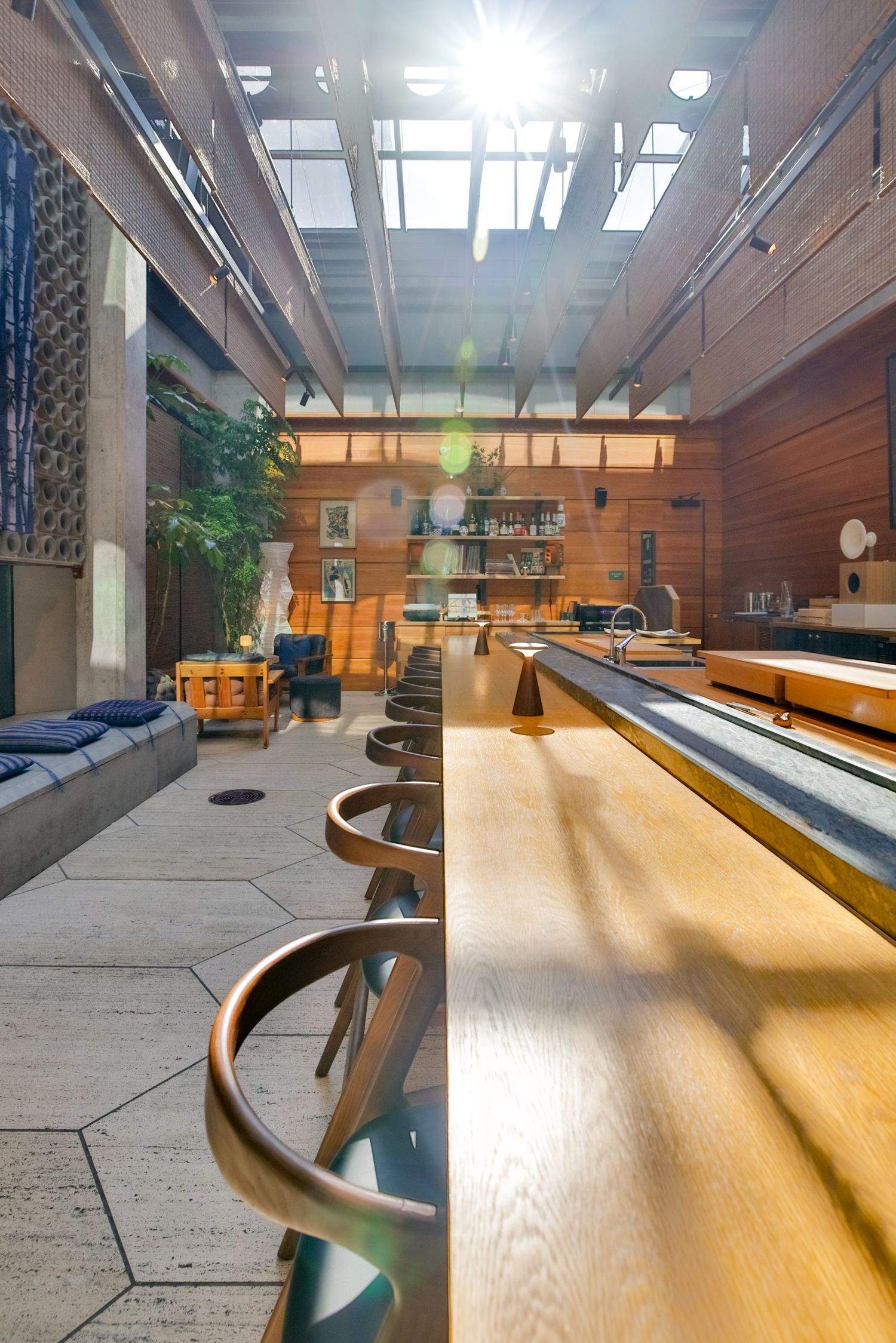 Sunlit modern bar interior with wooden counter, bar stools, skylights, and cozy lounge seating area.