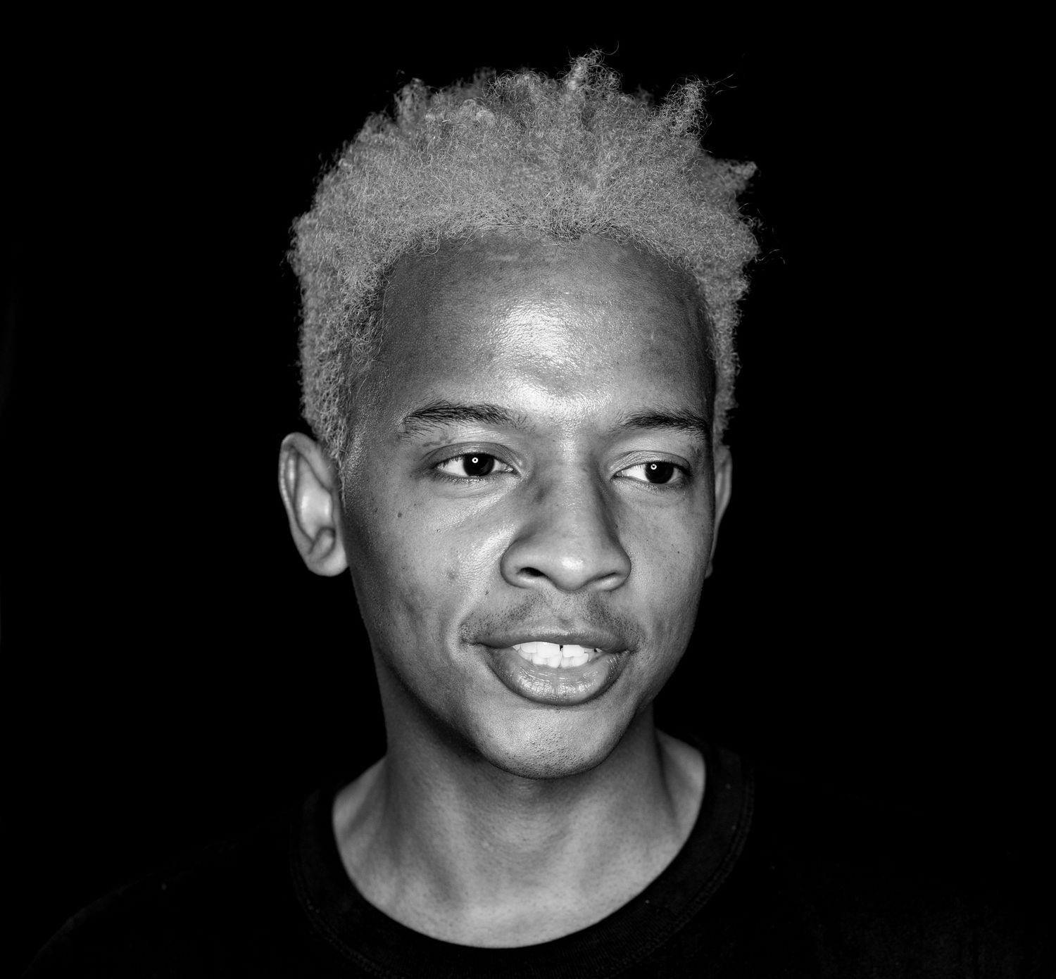 Black and white portrait of a young man with bleached hair against dark background.