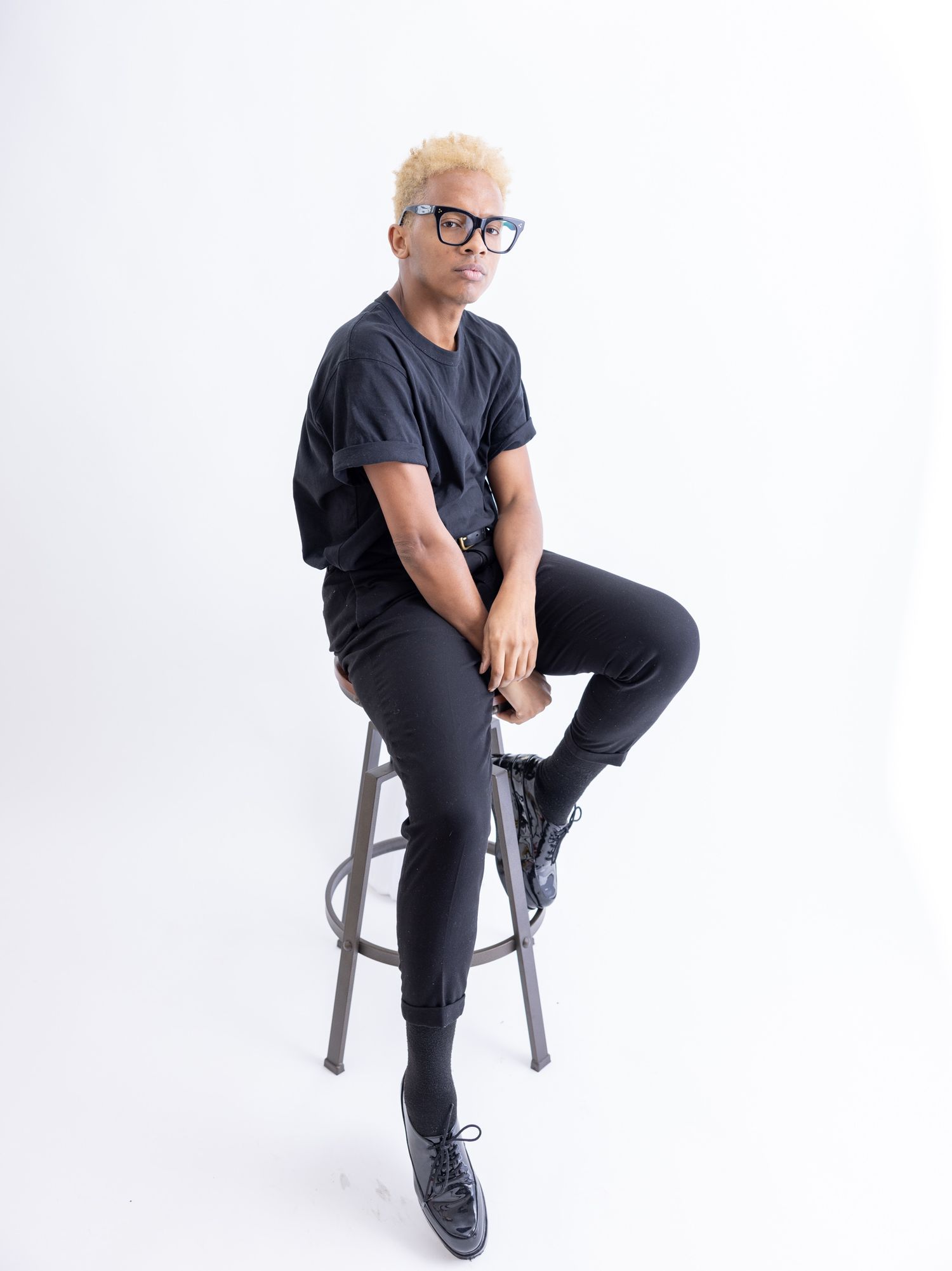 Person with blonde hair and glasses sitting on stool in all-black outfit on white background.