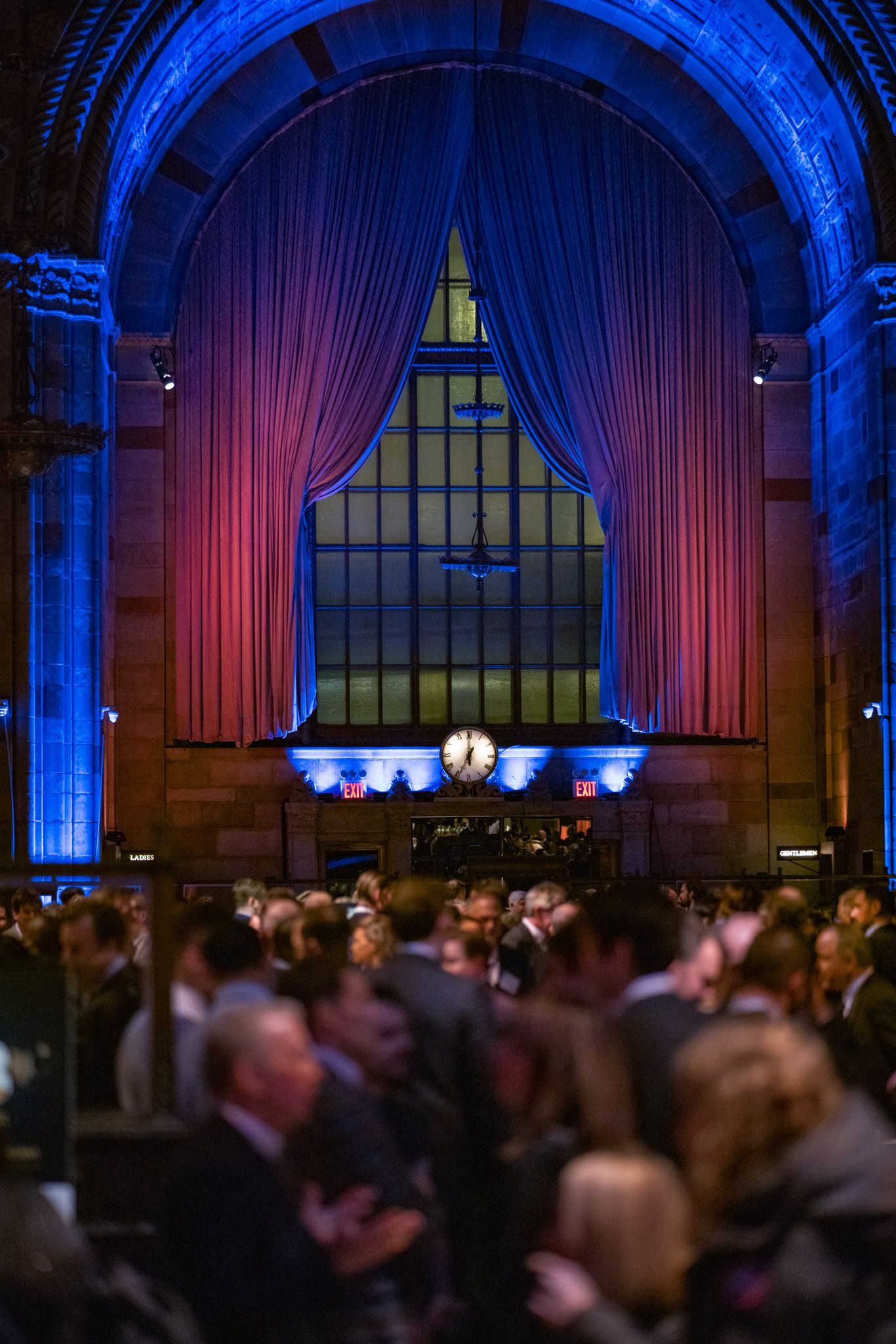 Crowded gala event inside historic venue with dramatic red curtains and blue arch lighting.