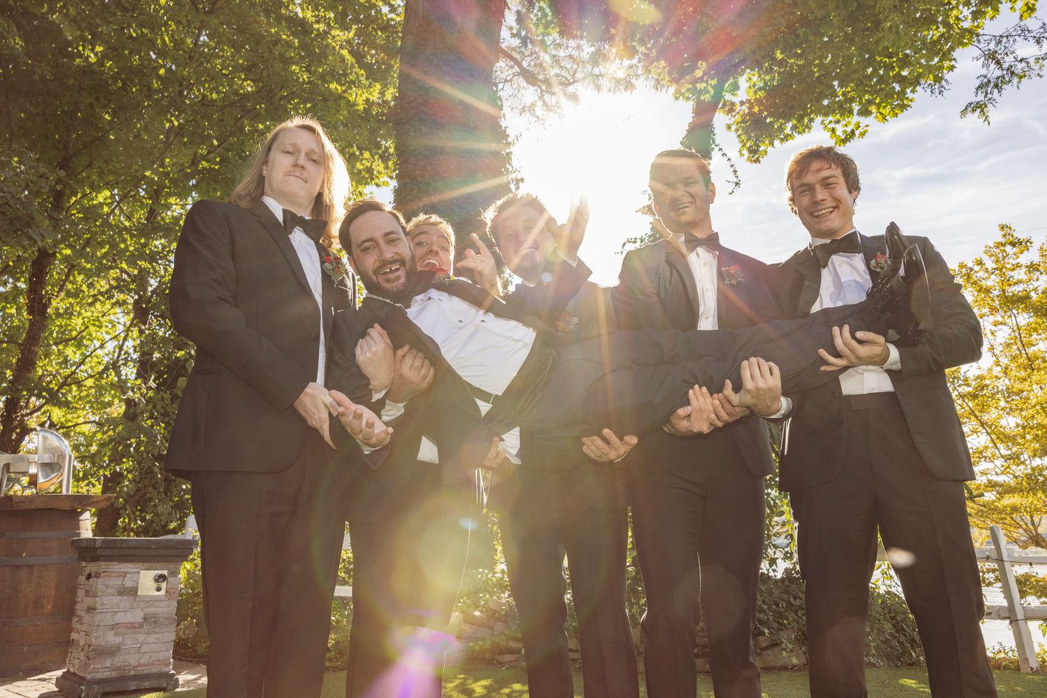 Groomsmen in suits celebrate outdoors with sunburst light effect filtering through trees.