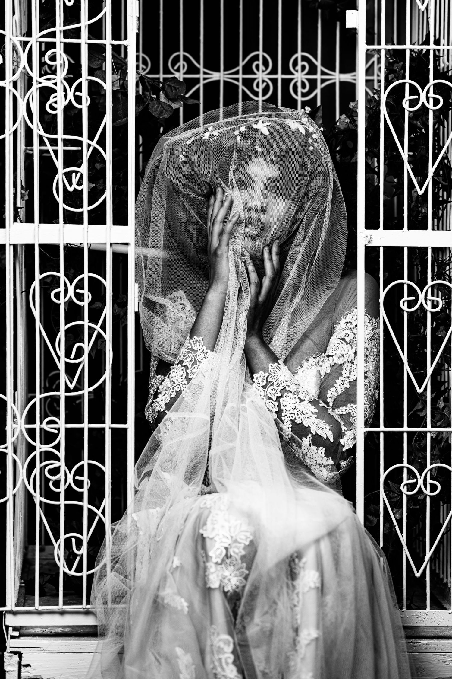 Black and white portrait of bride in lace wedding dress and veil praying by ornate gate.