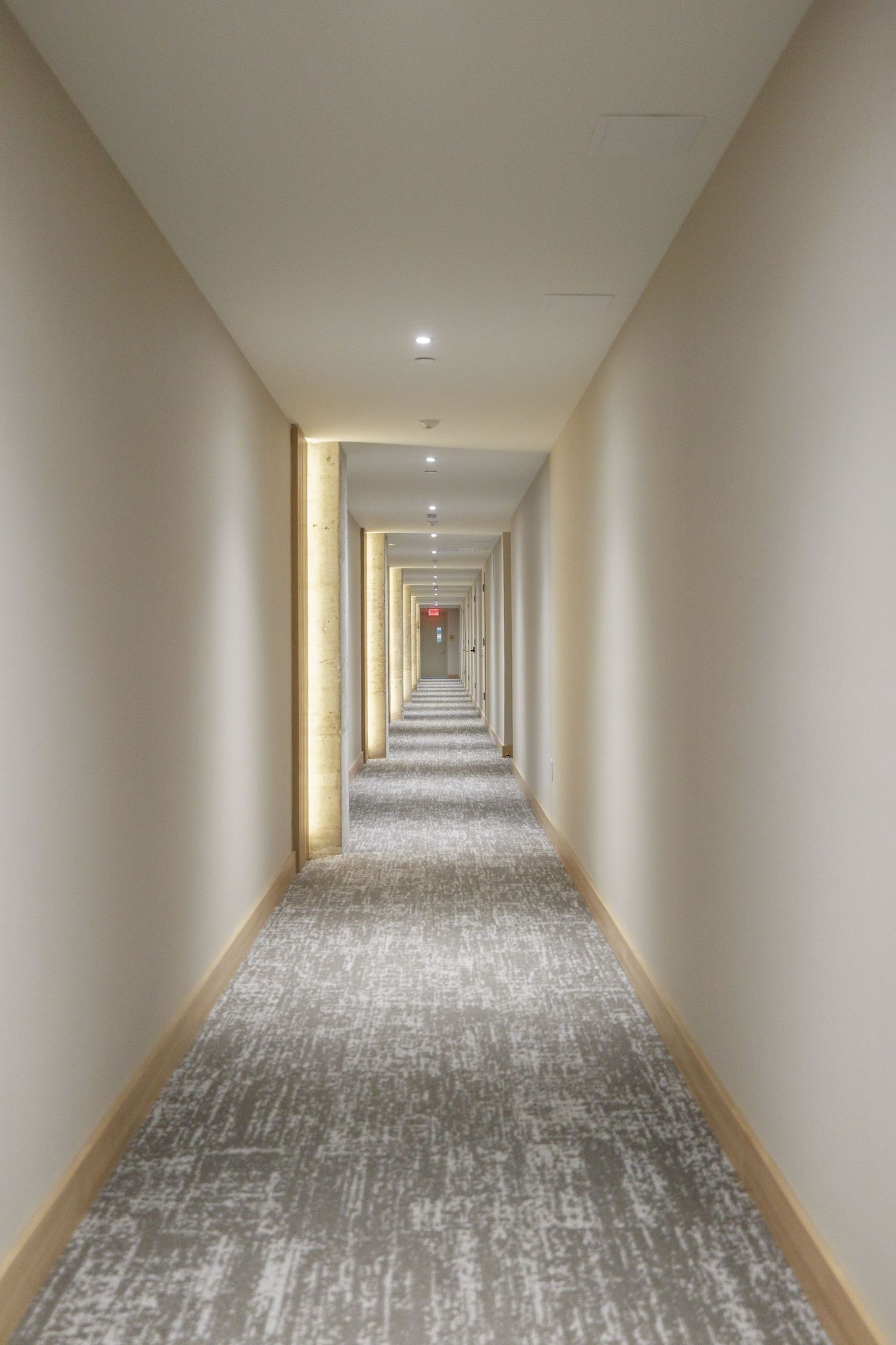 Long modern hotel corridor with patterned carpet, recessed lighting, and warm wood baseboards.