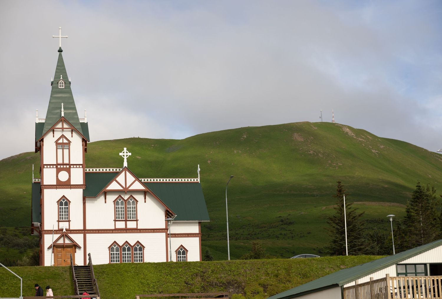 Traditional white wooden Norwegian-style church with green roof and steeple set against a lush green hillside.
