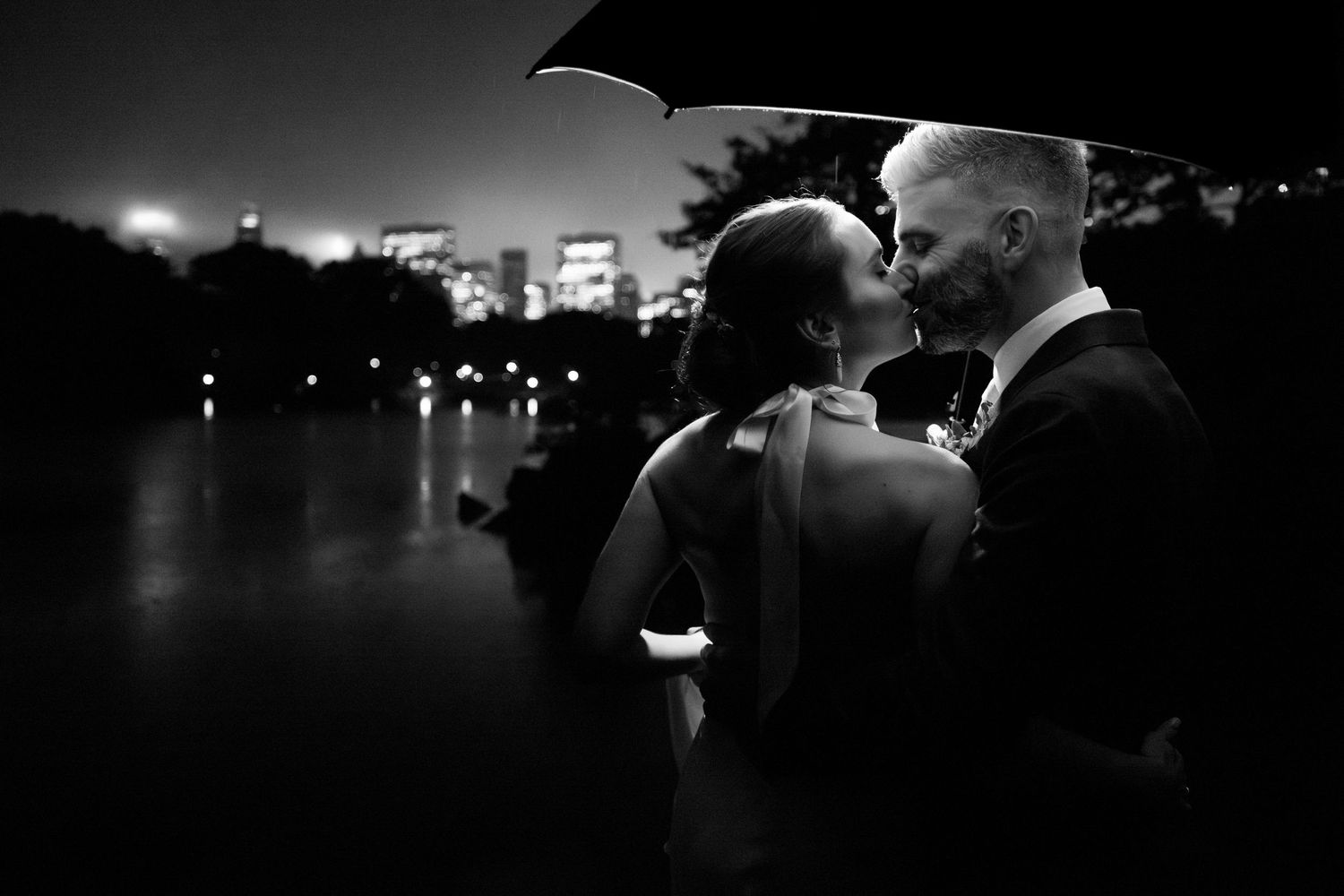 Bride and groom share a romantic kiss under an umbrella in a stunning black and white nighttime portrait.