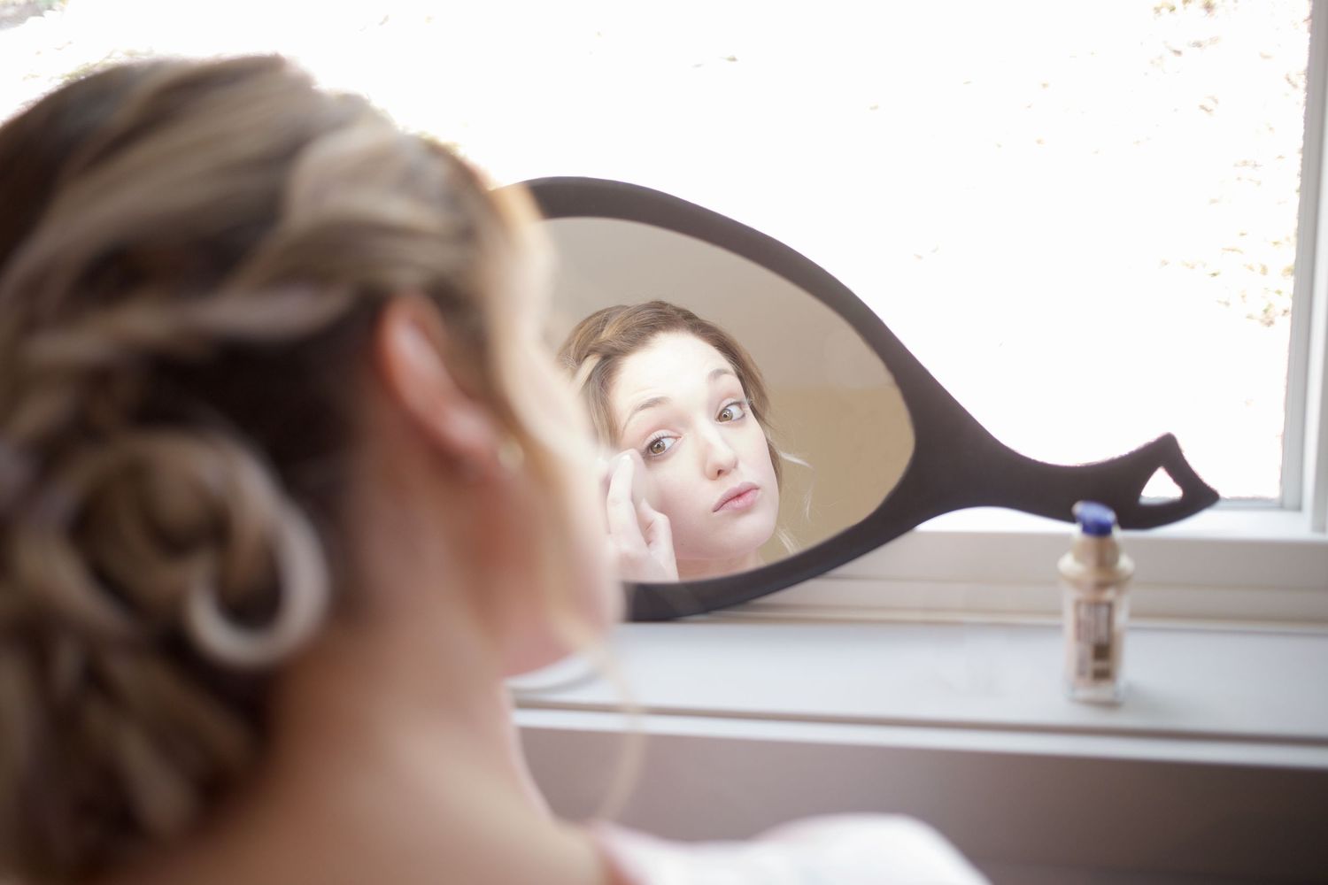 Bride checks her makeup in an oval hand mirror by a bright window before her wedding.
