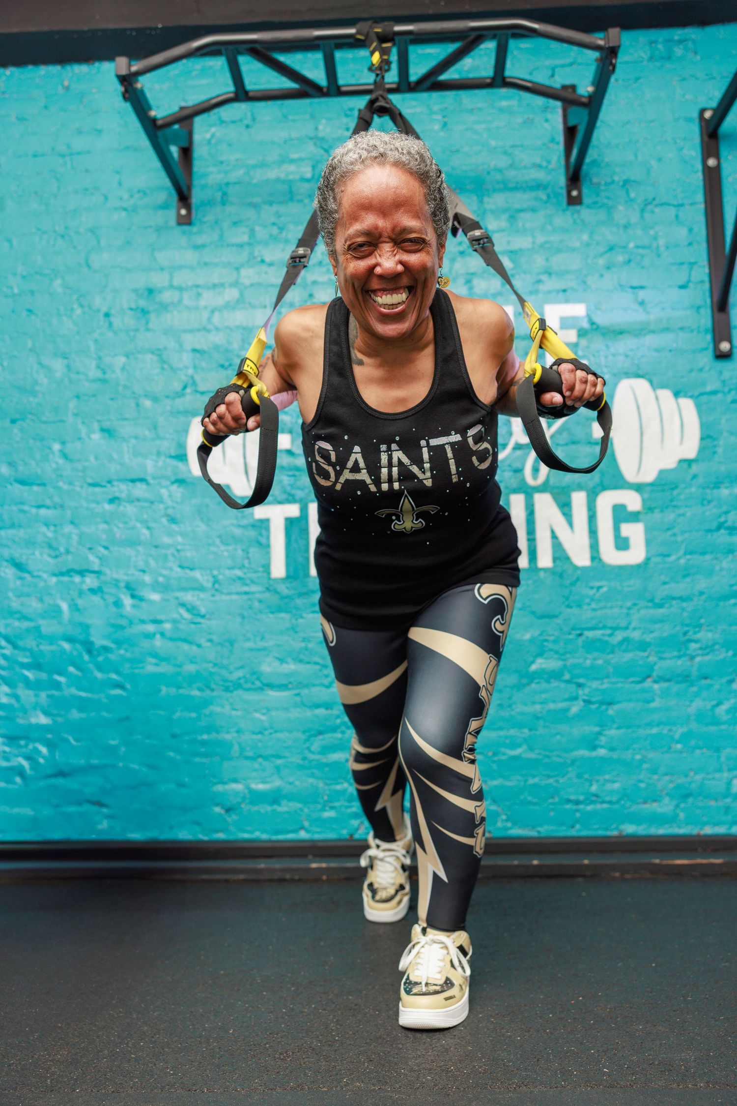 Energetic woman in Saints tank top laughing while using TRX straps in a vibrant teal gym.
