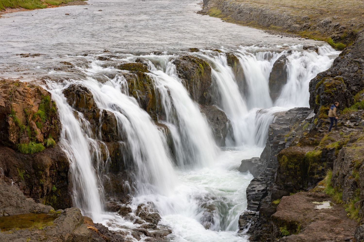 Multiple cascading waterfalls flowing over rocky terrain into a rushing river in Iceland.