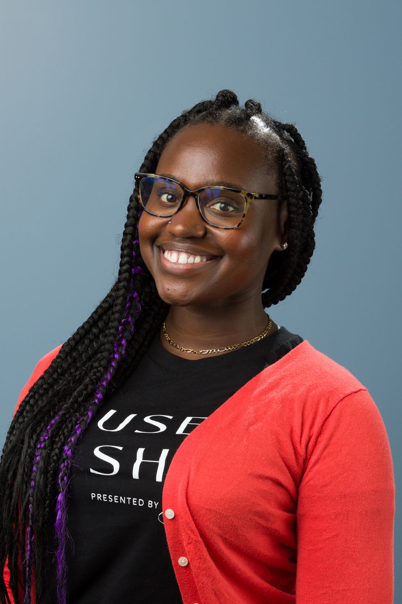 Smiling young Black woman with braided hair wearing glasses and red cardigan posing for a headshot.