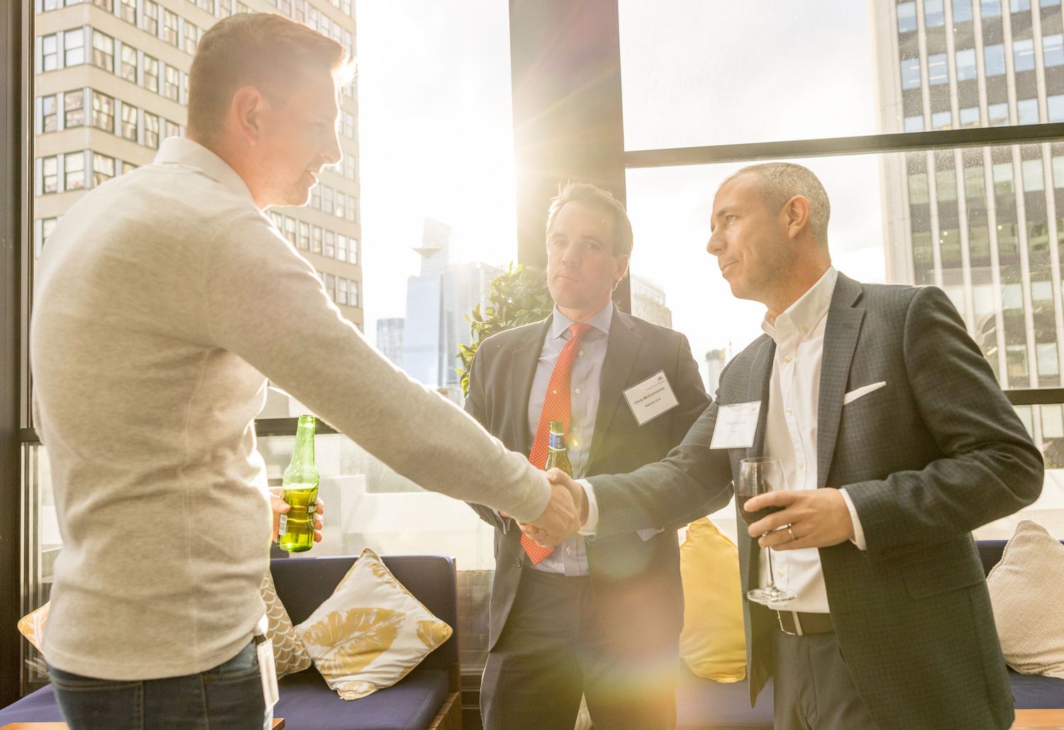 Three businessmen shaking hands and networking at a professional conference event with city skyline in background.