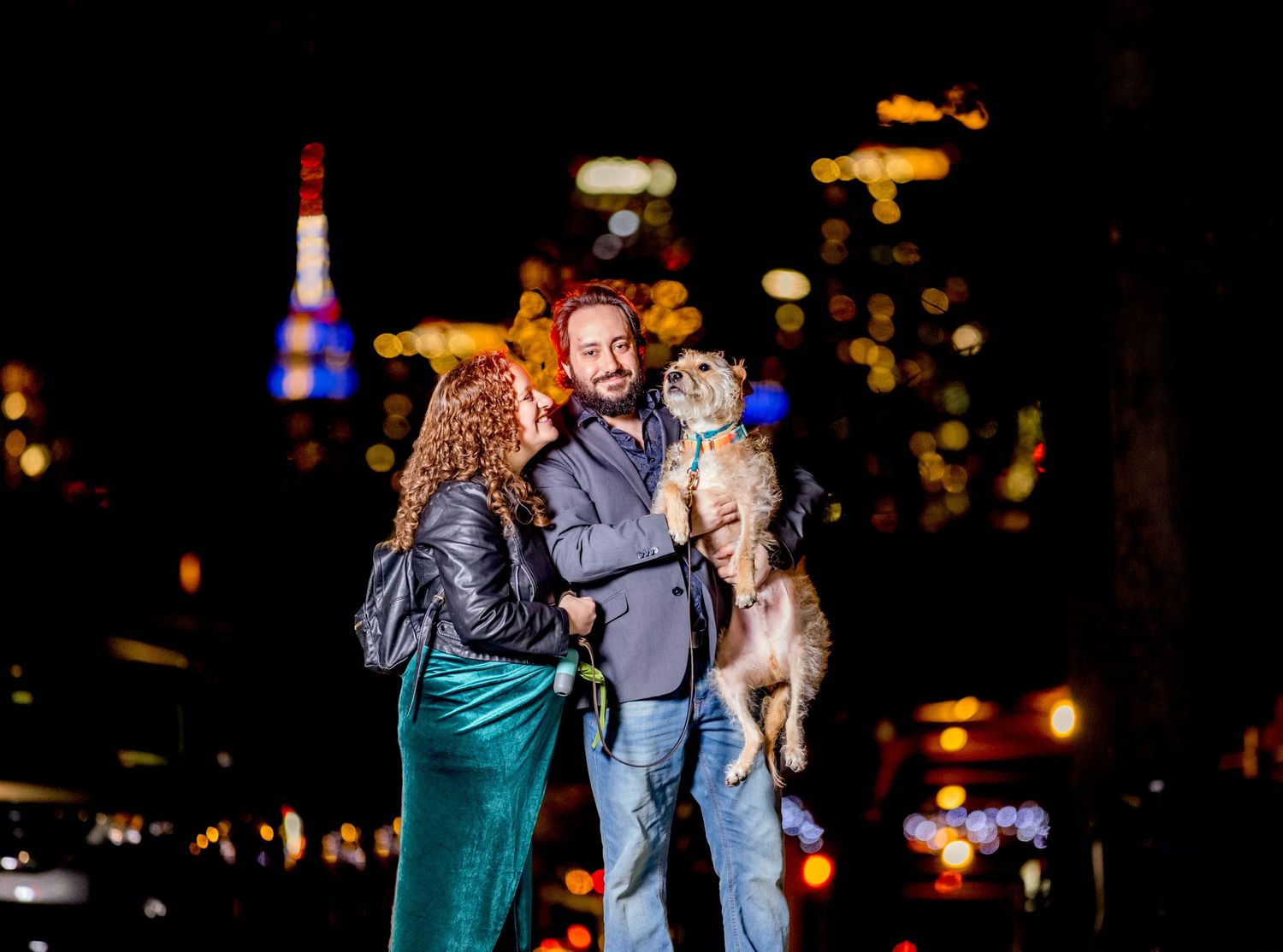 Couple holding a dog together at night with city lights bokeh in background.