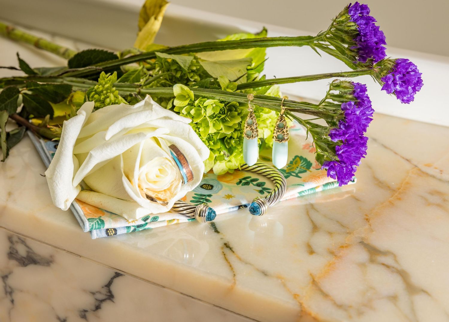 White rose and purple flowers arranged with turquoise jewelry on a marble surface.