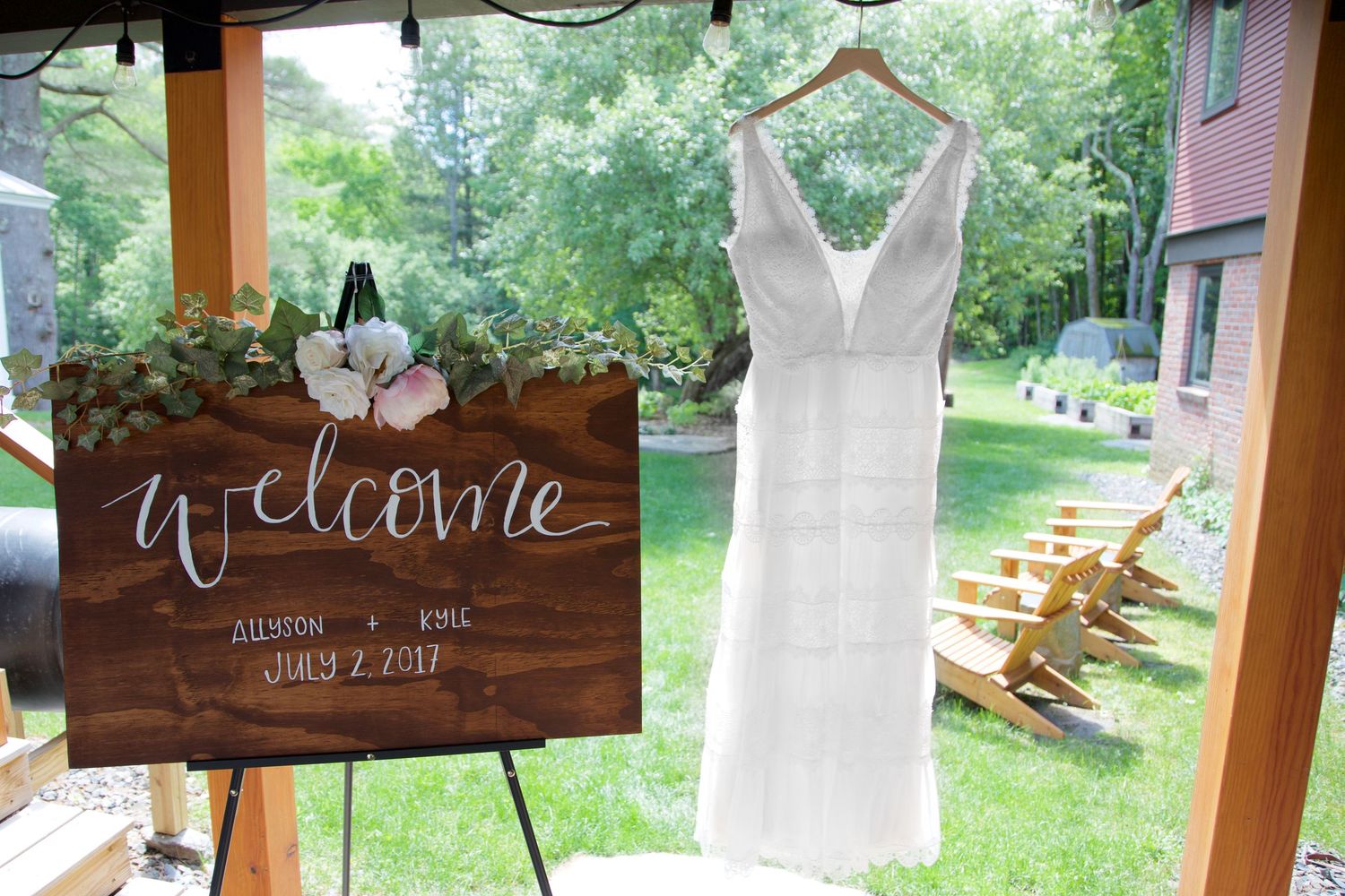 White wedding dress hanging beside a rustic wooden welcome sign at an outdoor summer wedding.