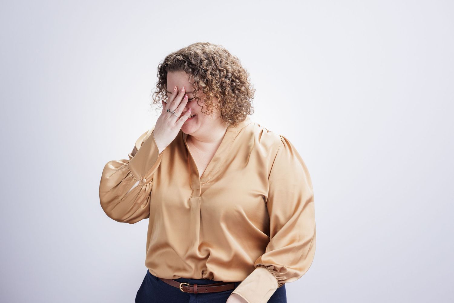 Curly-haired woman in gold satin blouse doing a facepalm gesture against white background.