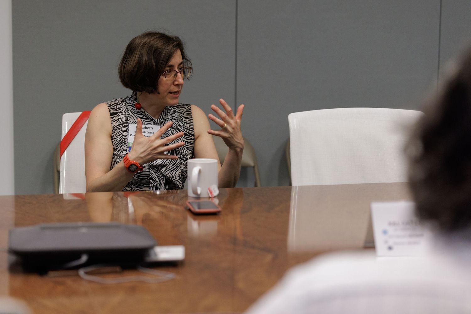A woman with short dark hair gestures expressively while speaking at a conference room table.