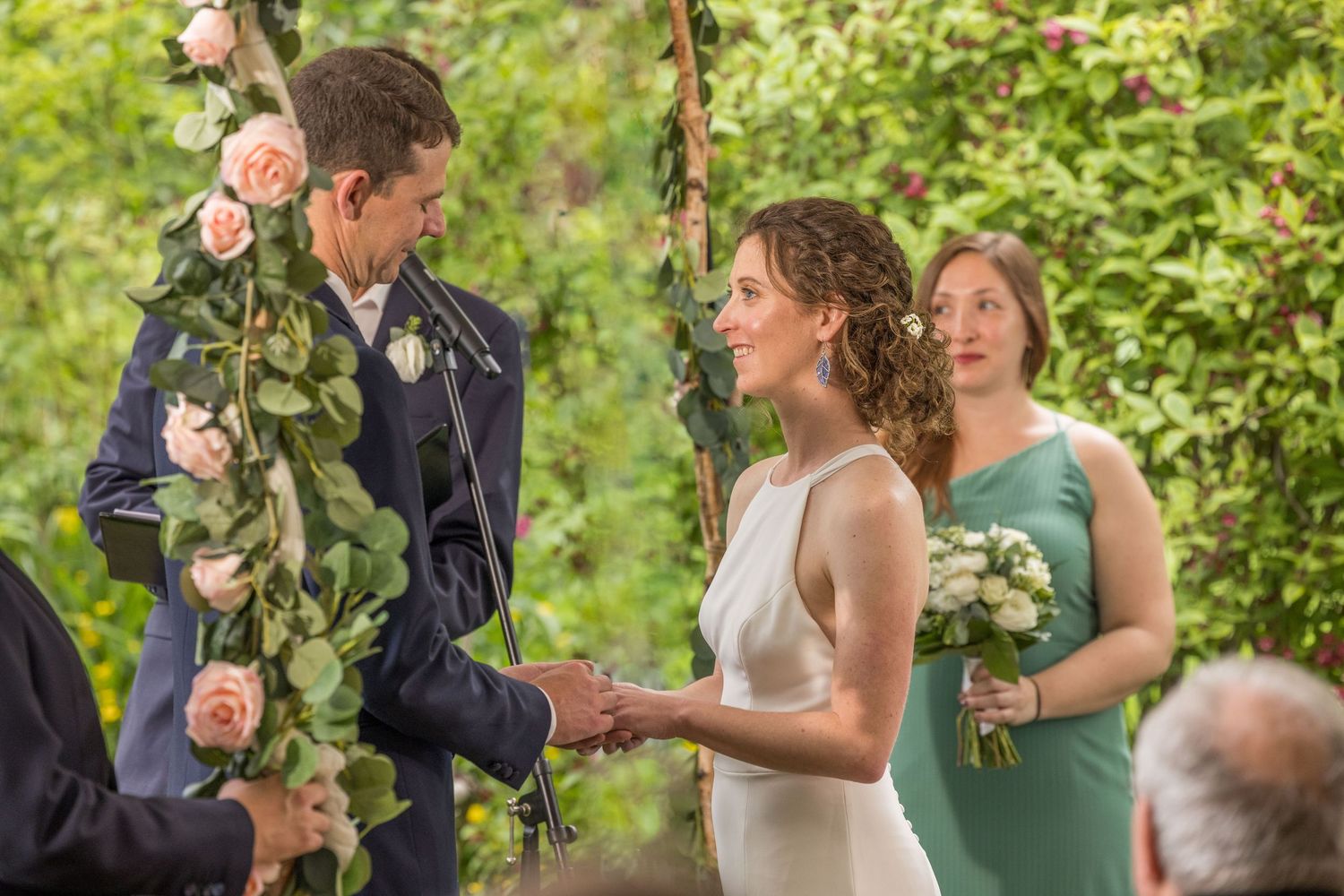 Bride and groom exchange vows outdoors under floral arch with pink roses and greenery at garden wedding.