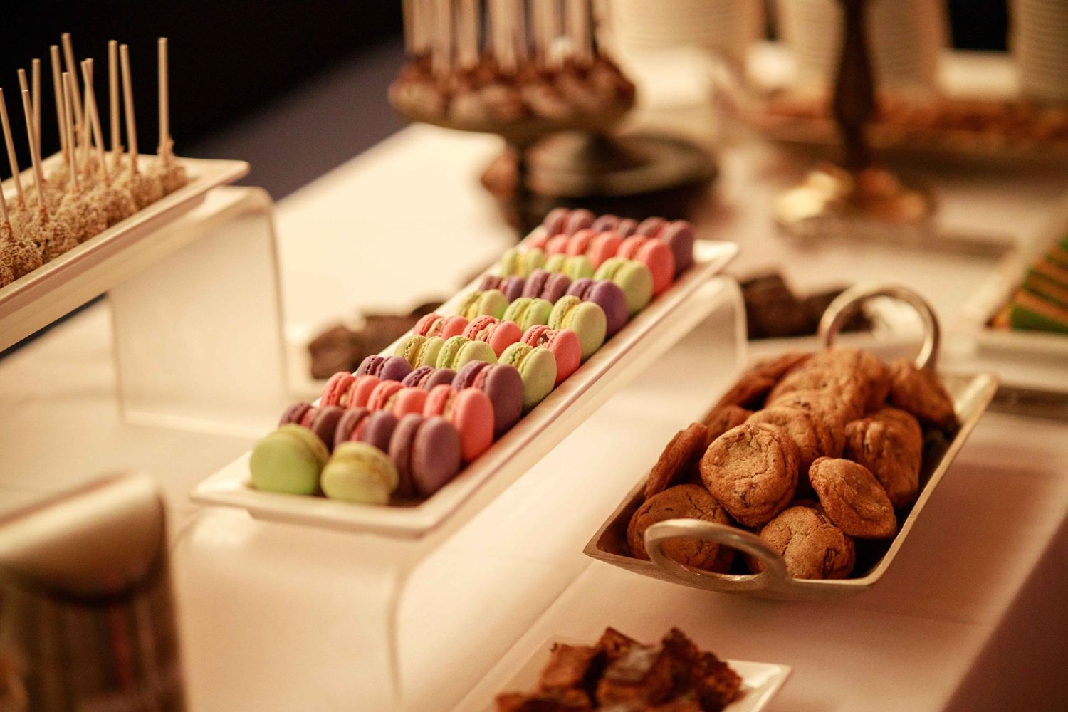 Dessert table display featuring colorful macarons on skewers and assorted pastries in white trays.