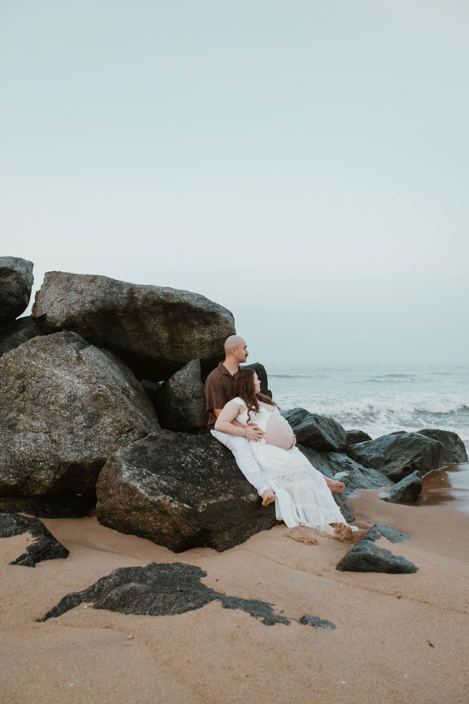 A couple sits together on coastal rocks at the beach, the woman in a white dress cradling a baby bump.