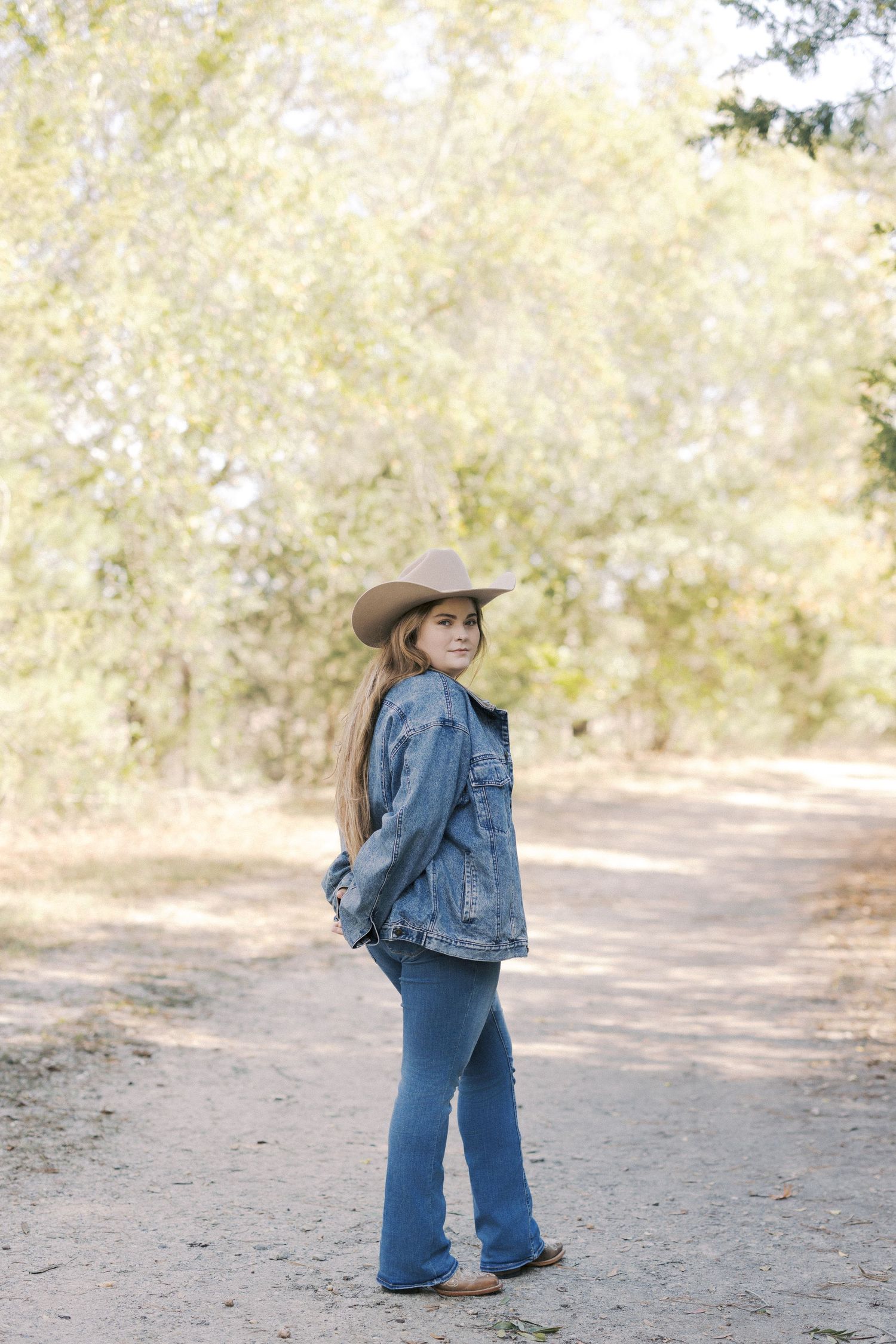 A person in a denim jacket and cowboy hat walks along a dirt path through a sun-dappled wooded area.