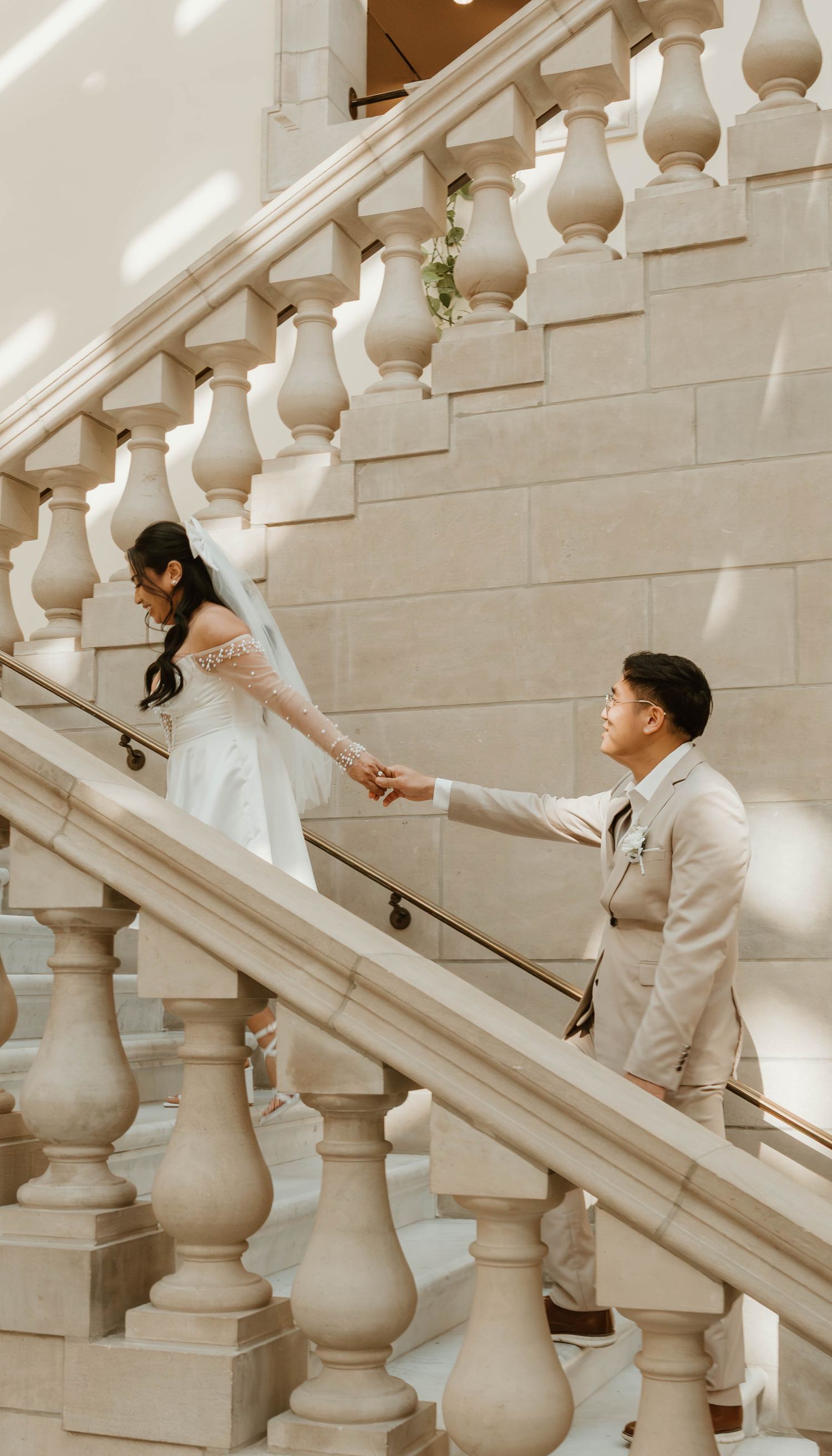 A bride and groom hold hands on an elegant stone staircase with ornate balusters during their wedding day.