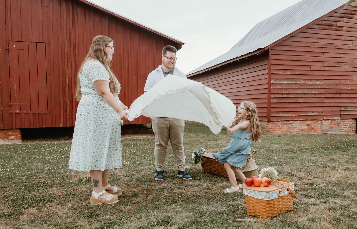 Family spreading a white blanket for a picnic near a red barn, with a wicker basket and a young girl sitting on the grass.