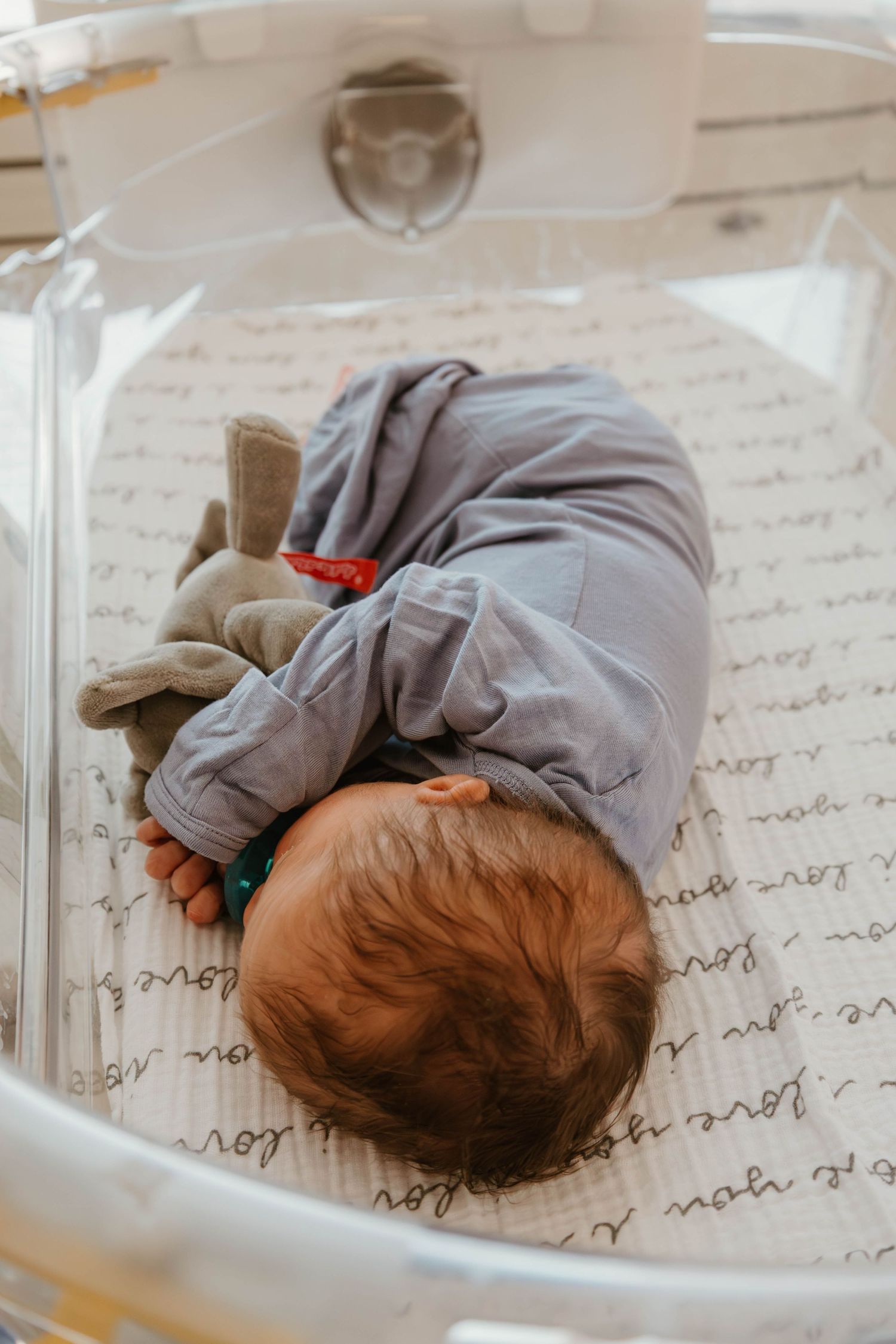 Overhead view of a sleeping infant in gray clothing lying on handwritten paper in a clear bassinet.