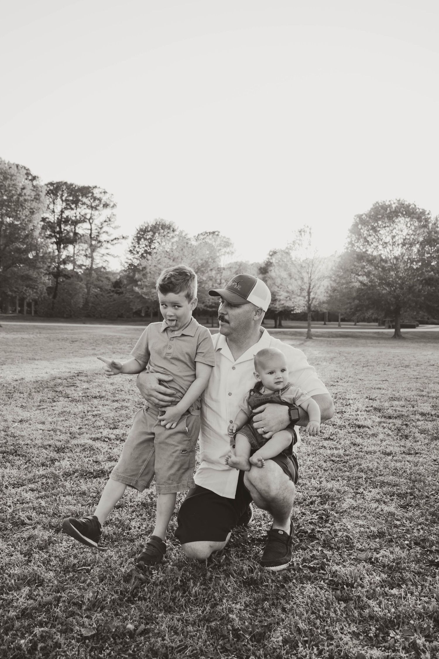 Father kneels on grass holding baby while young son stands beside him outdoors.