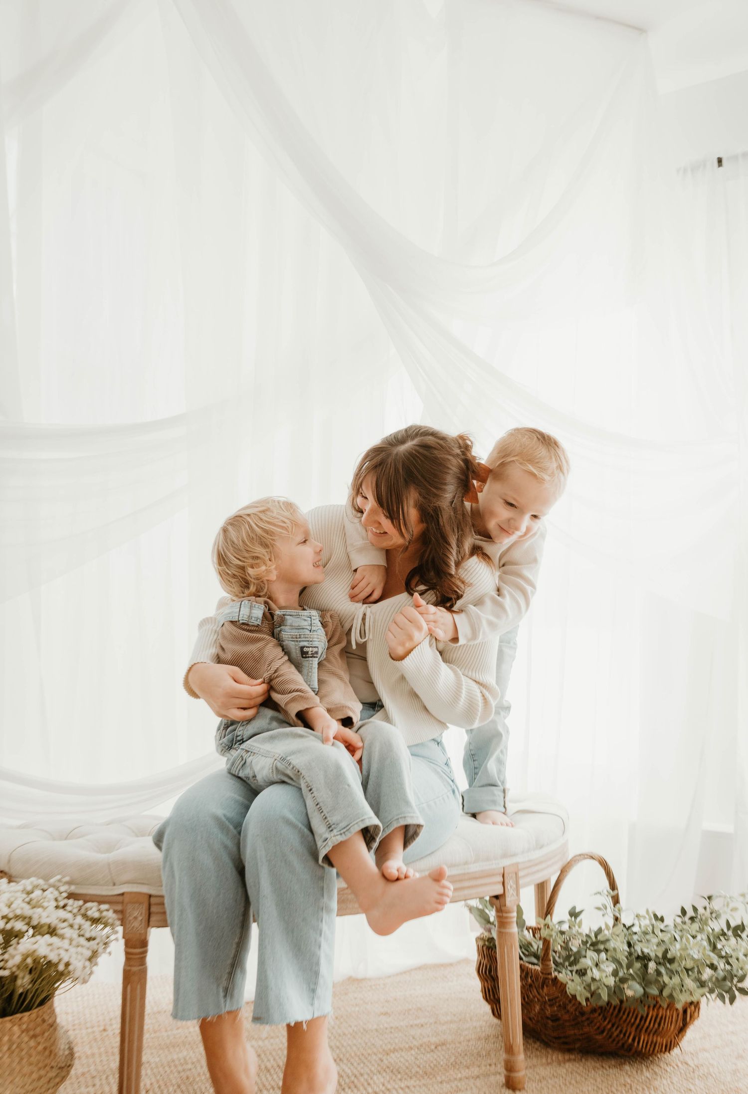 Three siblings in matching blue outfits share sweet hugs and kisses while sitting on a bench in a bright white room.