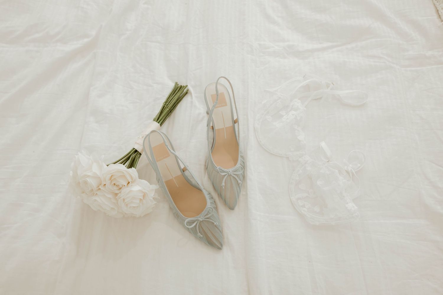 Elegant silver slingback heels with bow detail beside white roses bouquet and lace garter on white fabric.
