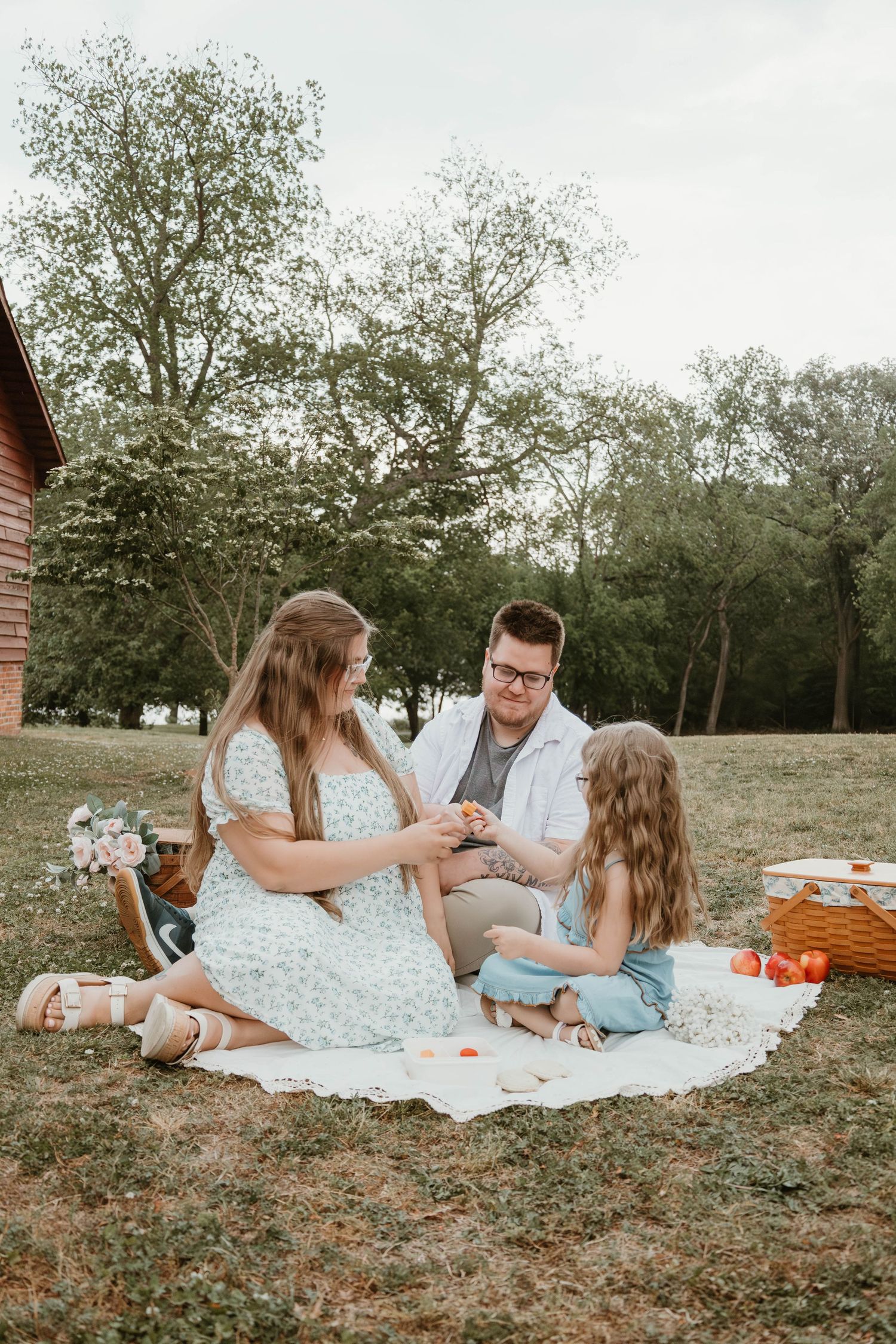 A family of three enjoys a cozy outdoor picnic on a blanket surrounded by lush green trees and a wicker basket.