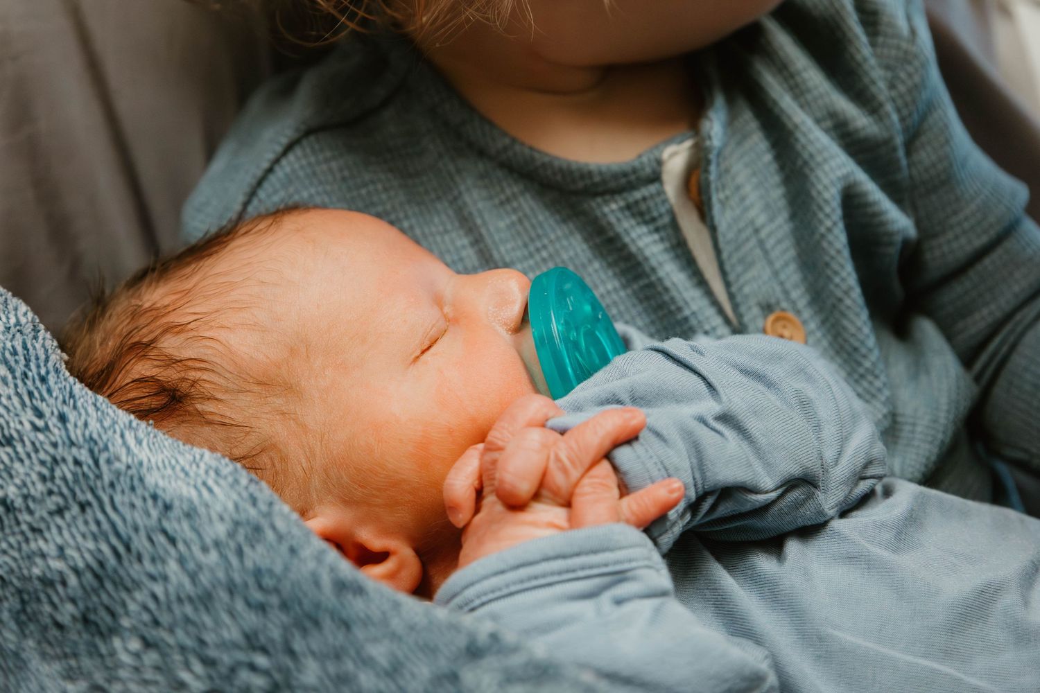 Close-up view of an infant sleeping soundly with a turquoise pacifier against gray fabric.