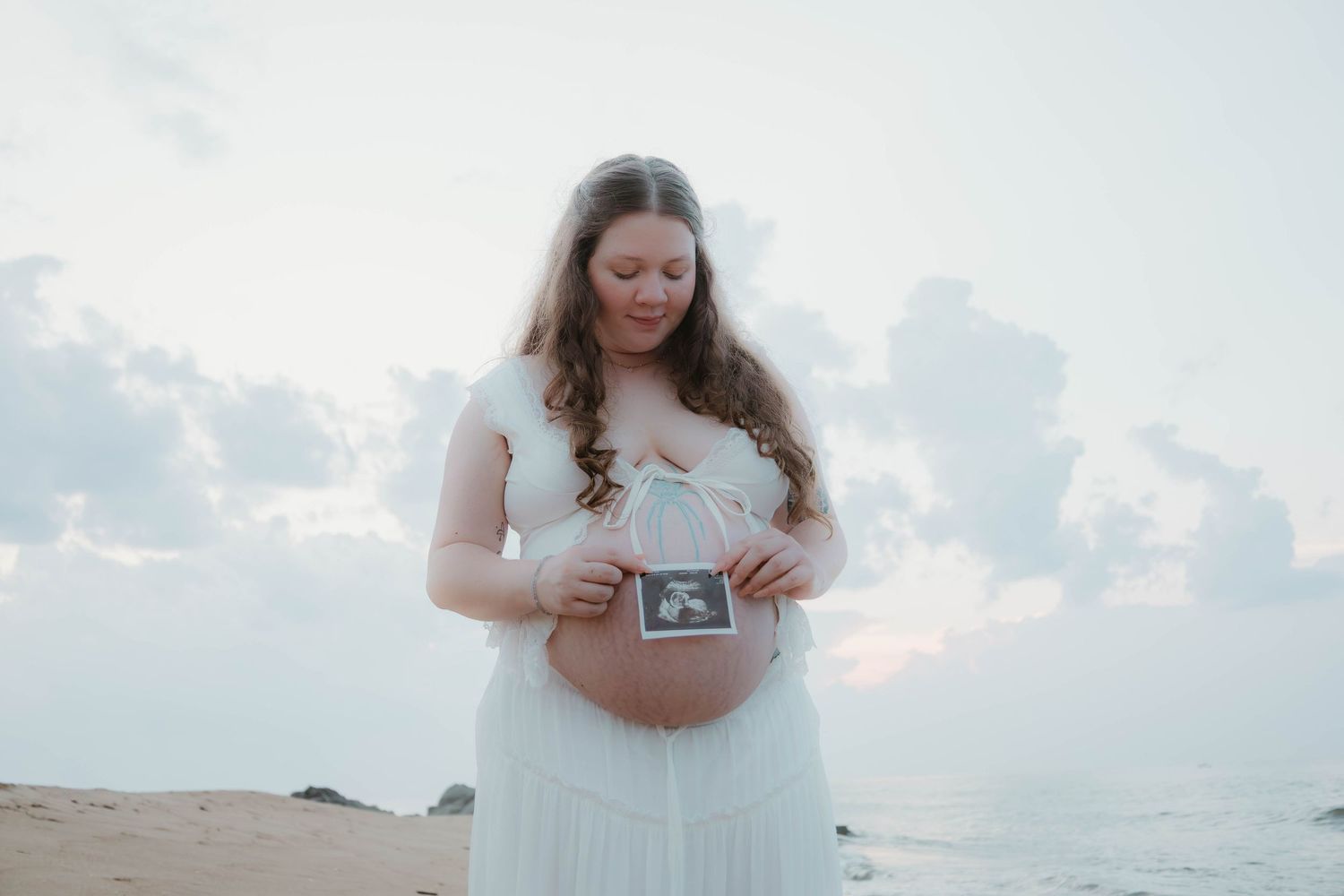 Pregnant woman in white dress holding ultrasound photo on beach at sunset.