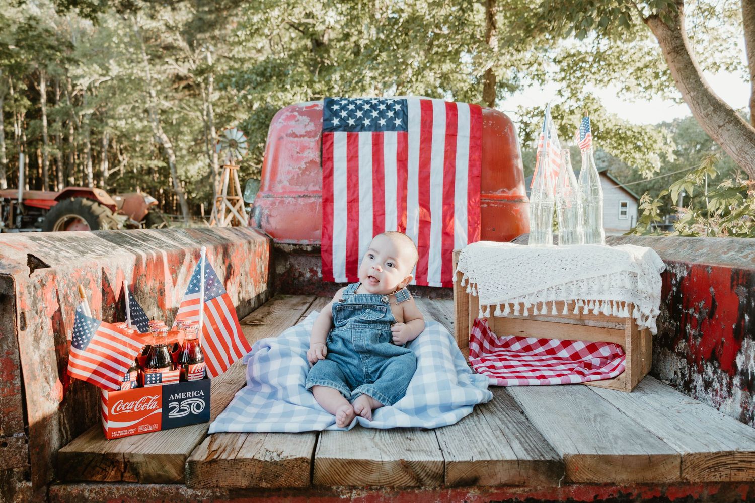 Baby in denim overalls sits on wooden deck with American flags and Coca-Cola bottles for a patriotic 4th of July photo.