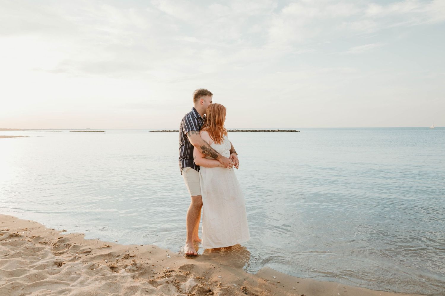 Couple embracing and kissing at the shoreline on a sandy beach at sunset, woman in white dress.