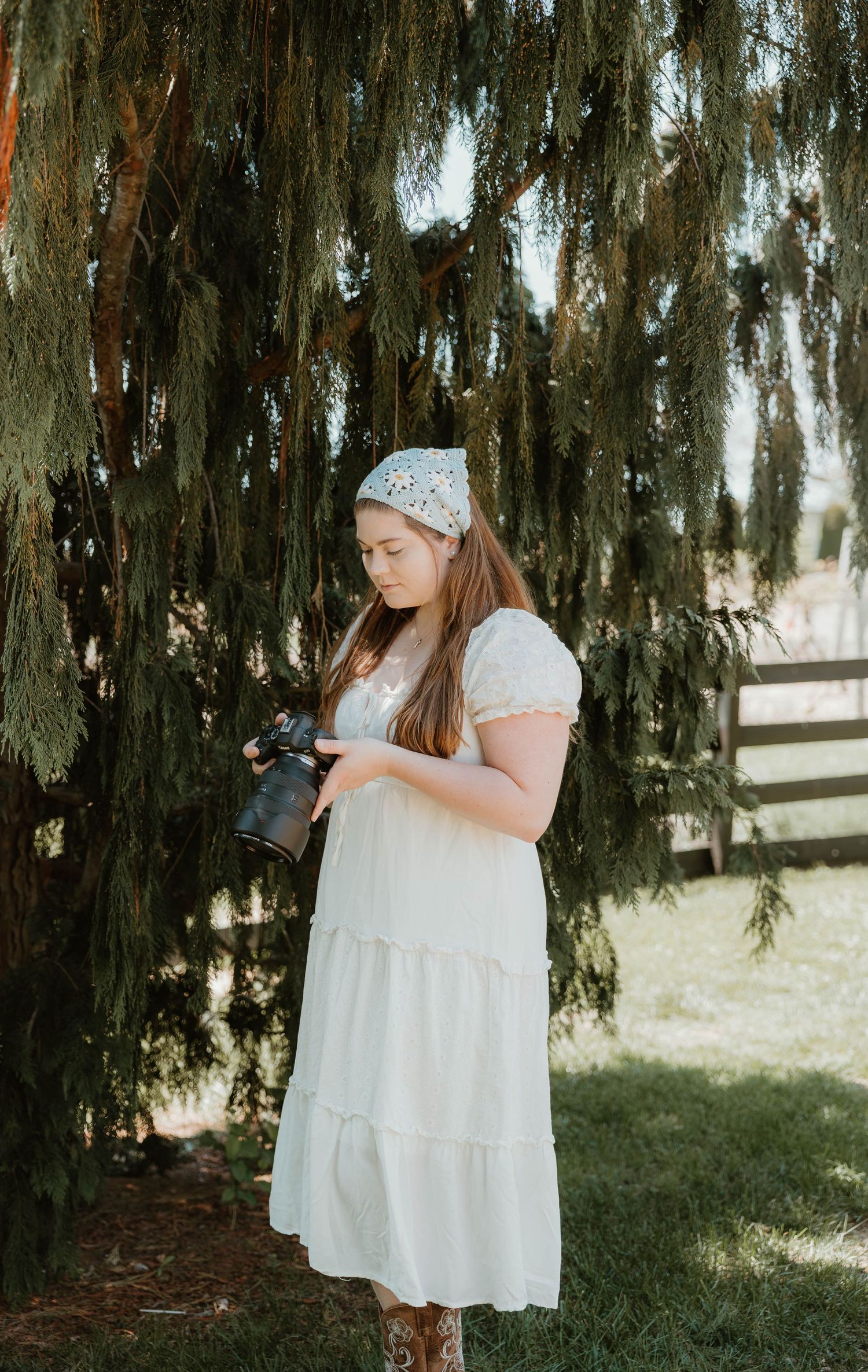 Young woman in white midi dress holding camera under weeping evergreen tree outdoors.