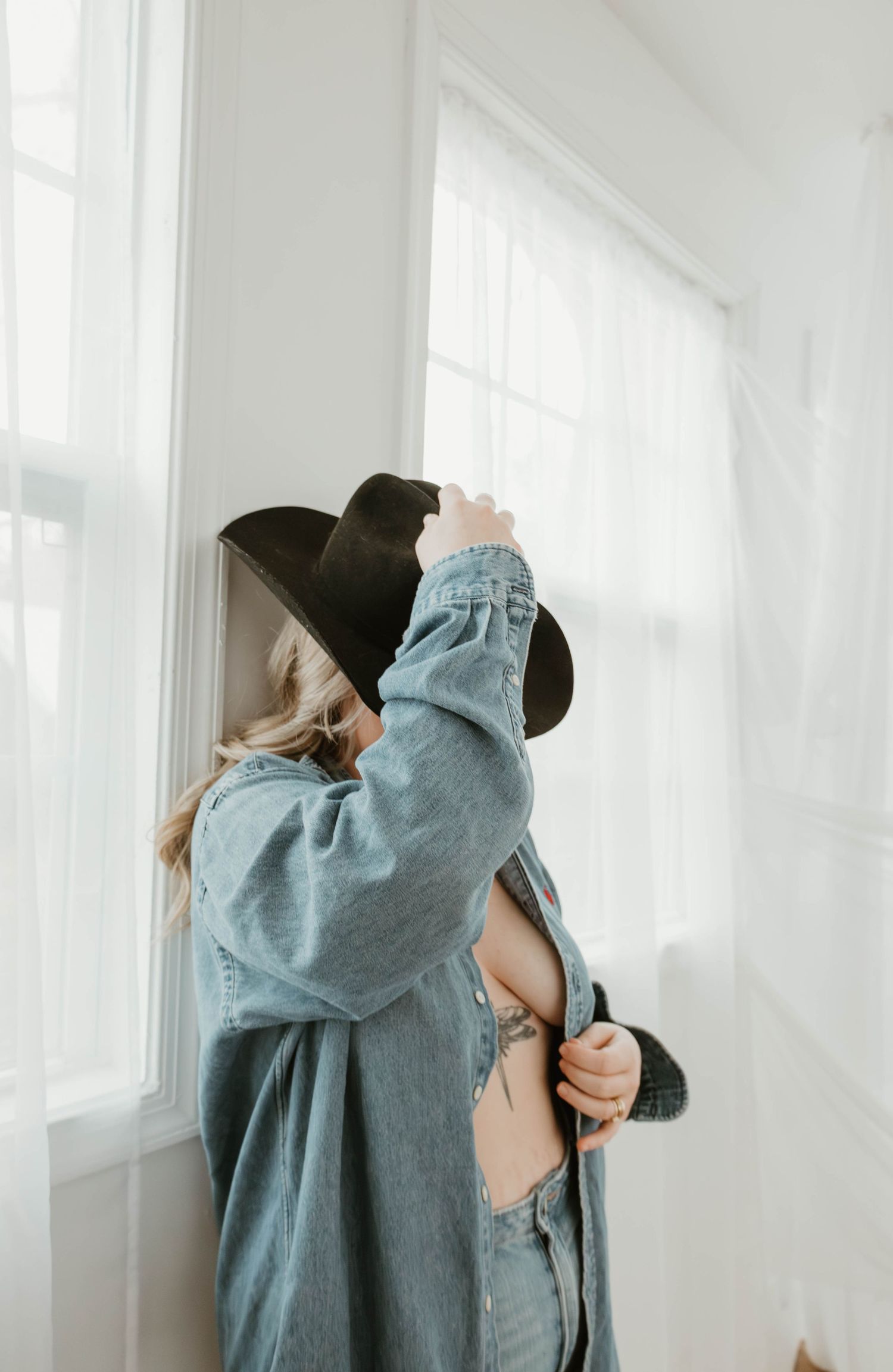 Woman in black hat and denim shirt standing by window in white room with cowboy boots.