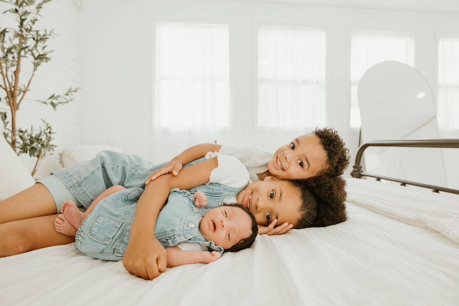 Three young siblings in denim outfits cuddle together on a white bed in a bright, airy bedroom.