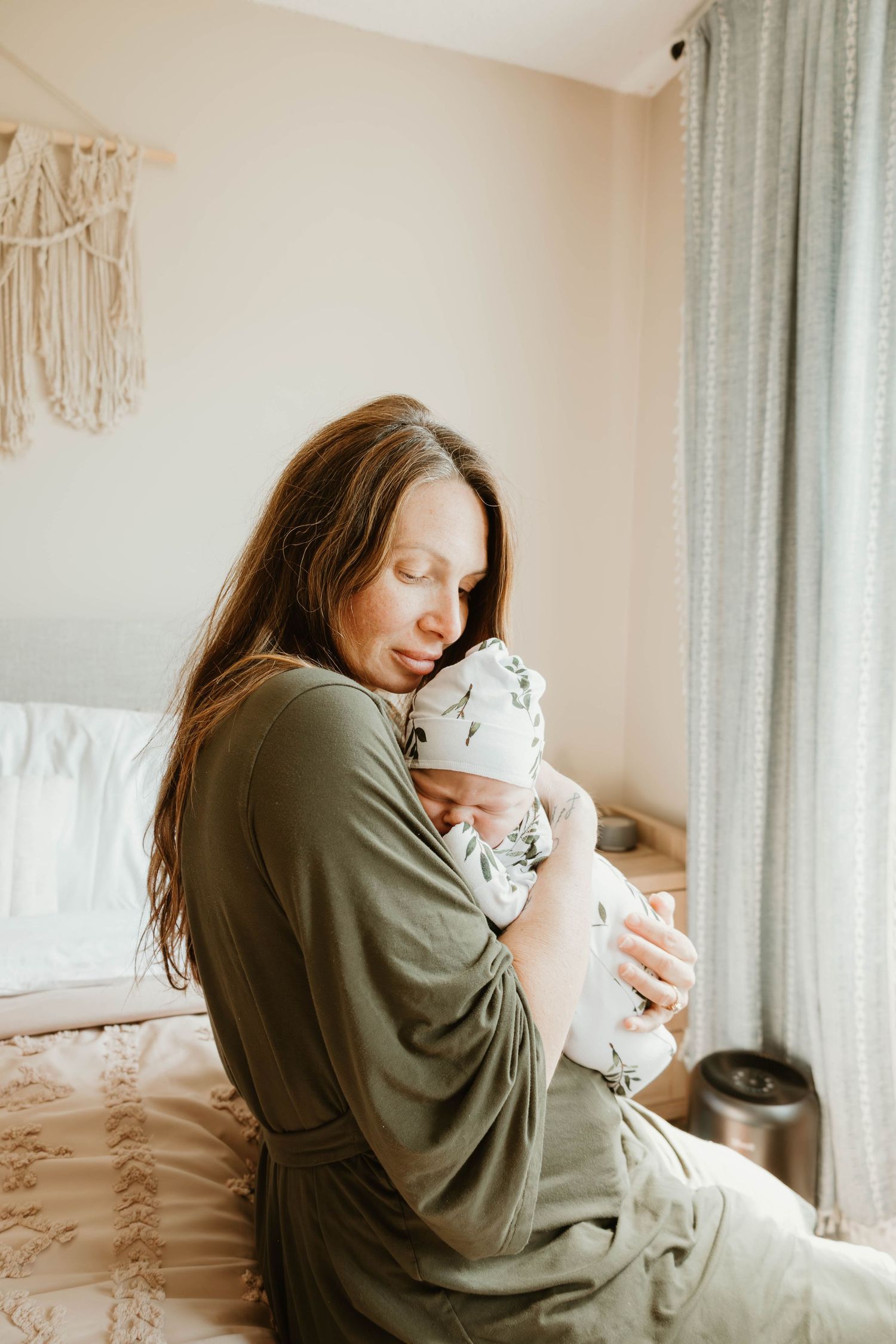 A mother in an olive green robe tenderly holds her newborn baby while sitting on a bed in a softly lit bedroom.